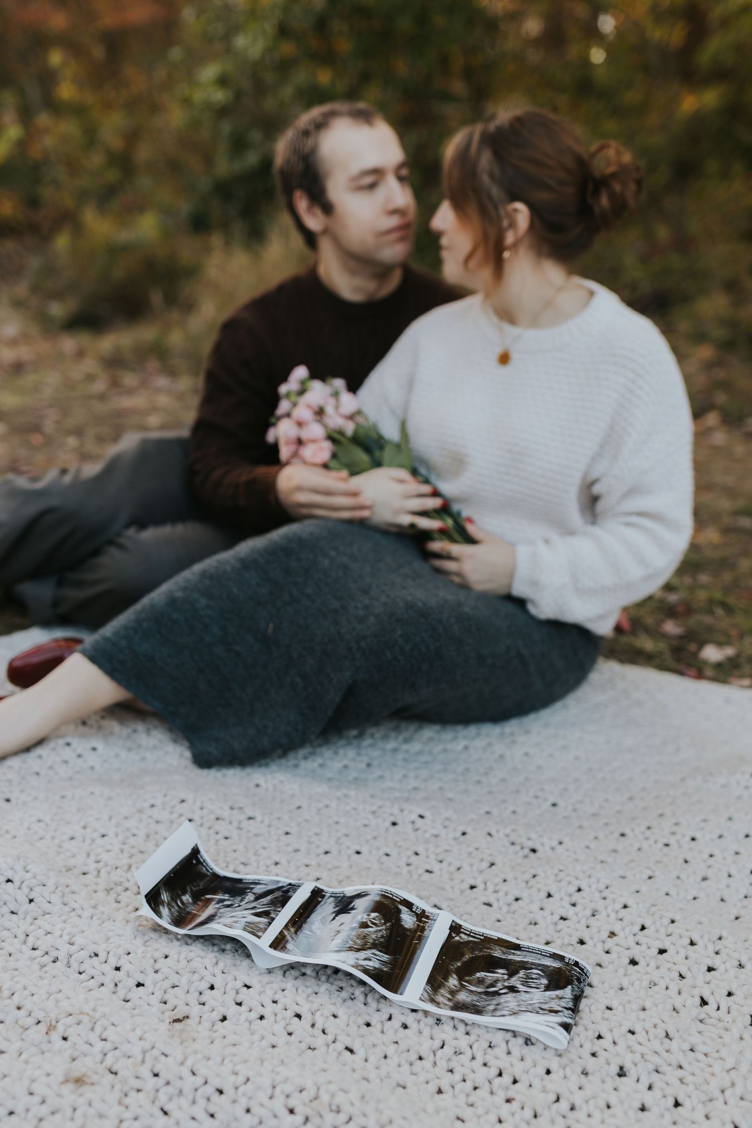 Couple sitting on blanket outdoors with ultrasound photos in foreground, pregnancy announcement photo.