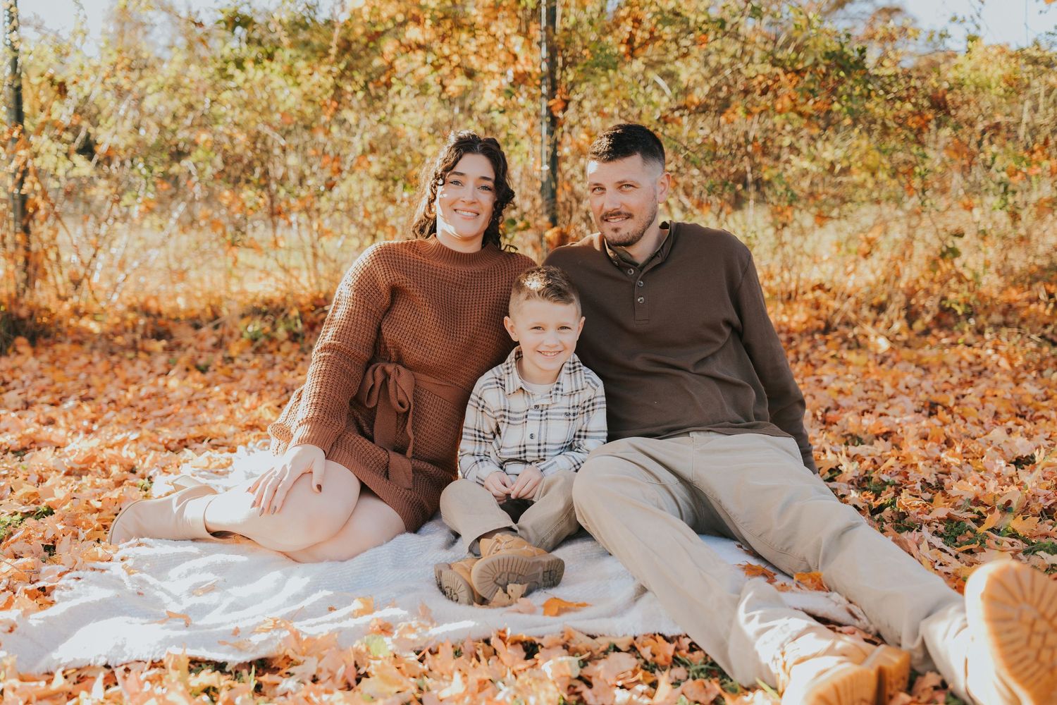 A happy family of three sits together on a blanket surrounded by colorful autumn foliage.