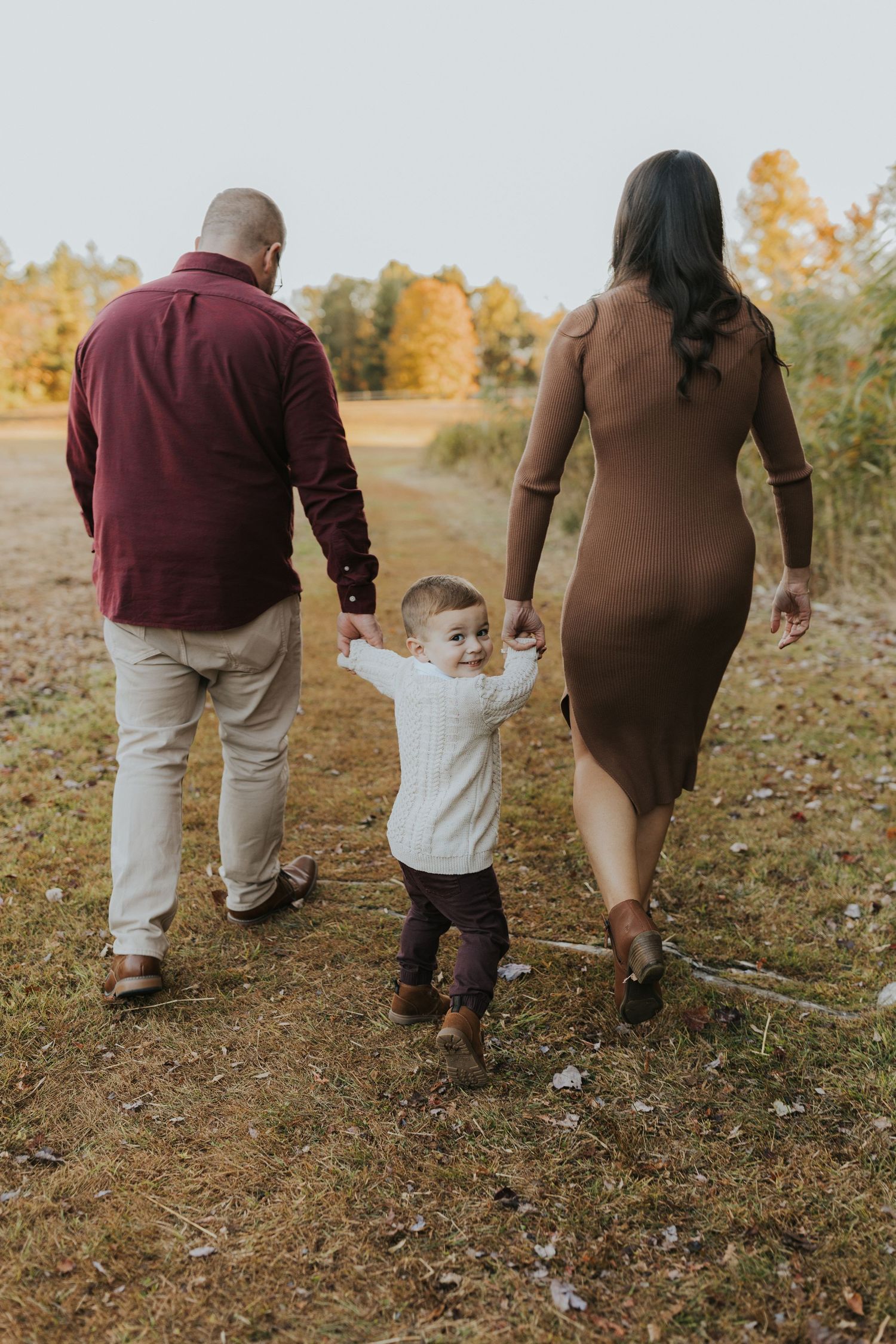 Family of three walking hand in hand down autumn path, toddler looking back at camera smiling.