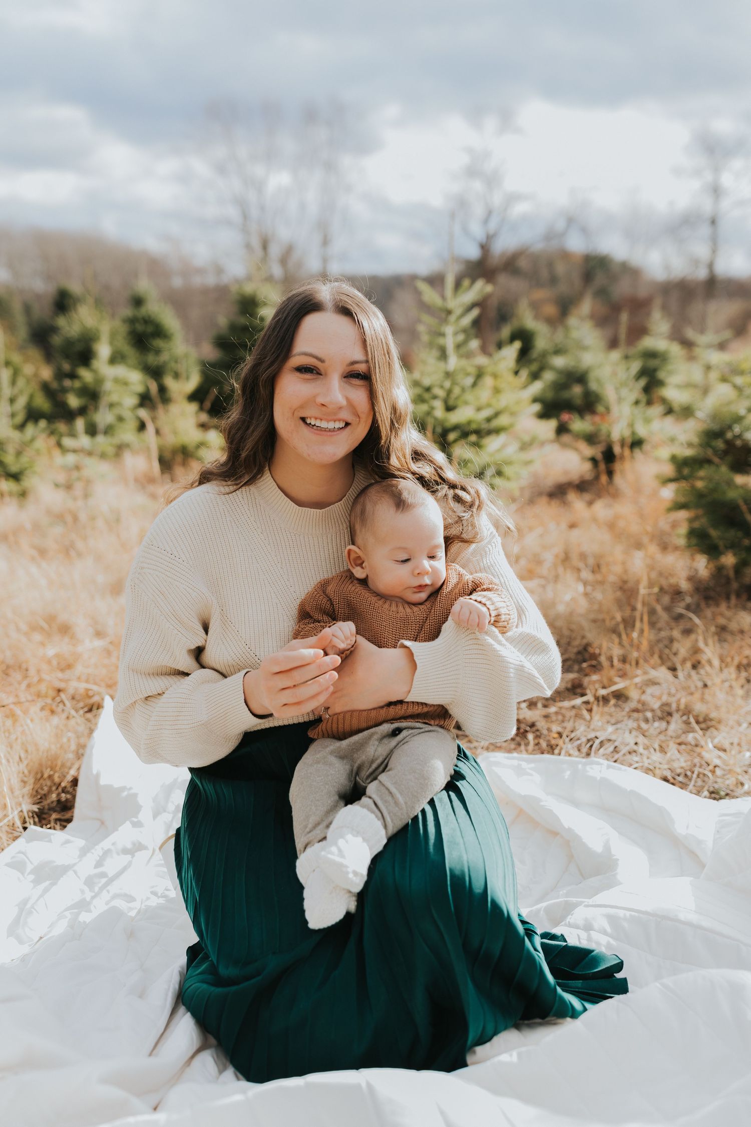 Smiling mother in cream sweater and green skirt holds baby on white blanket at tree farm.