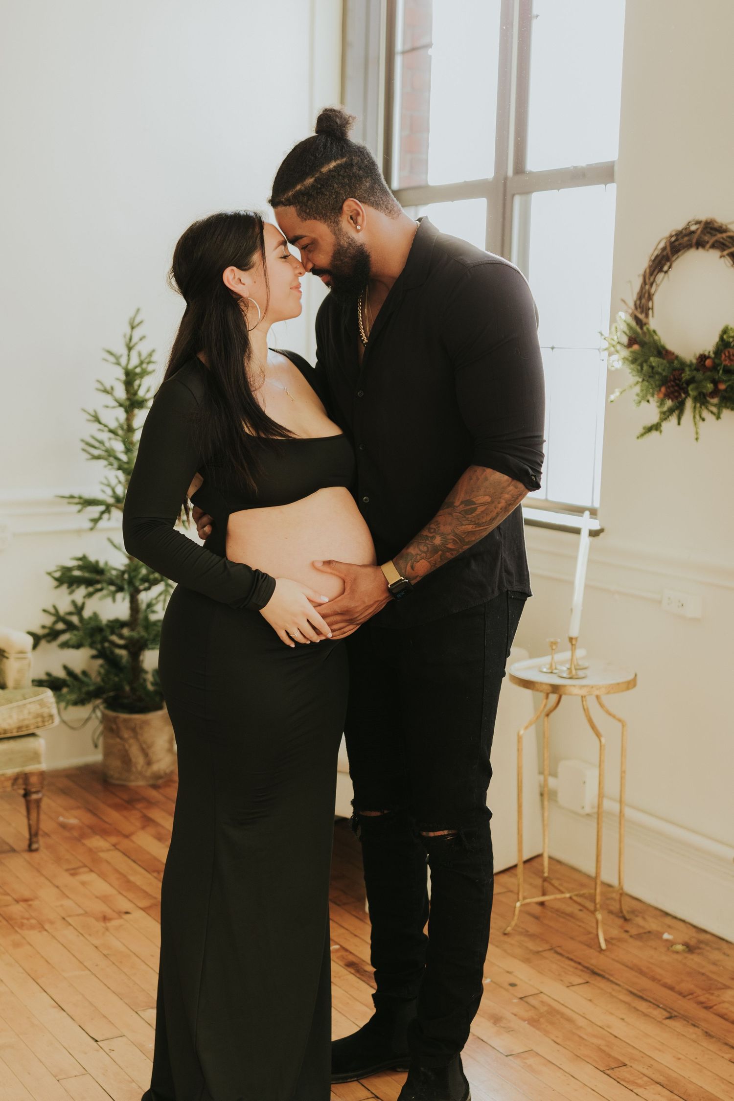 Couple in black sharing a kiss while man holds pregnant woman's belly near Christmas tree.