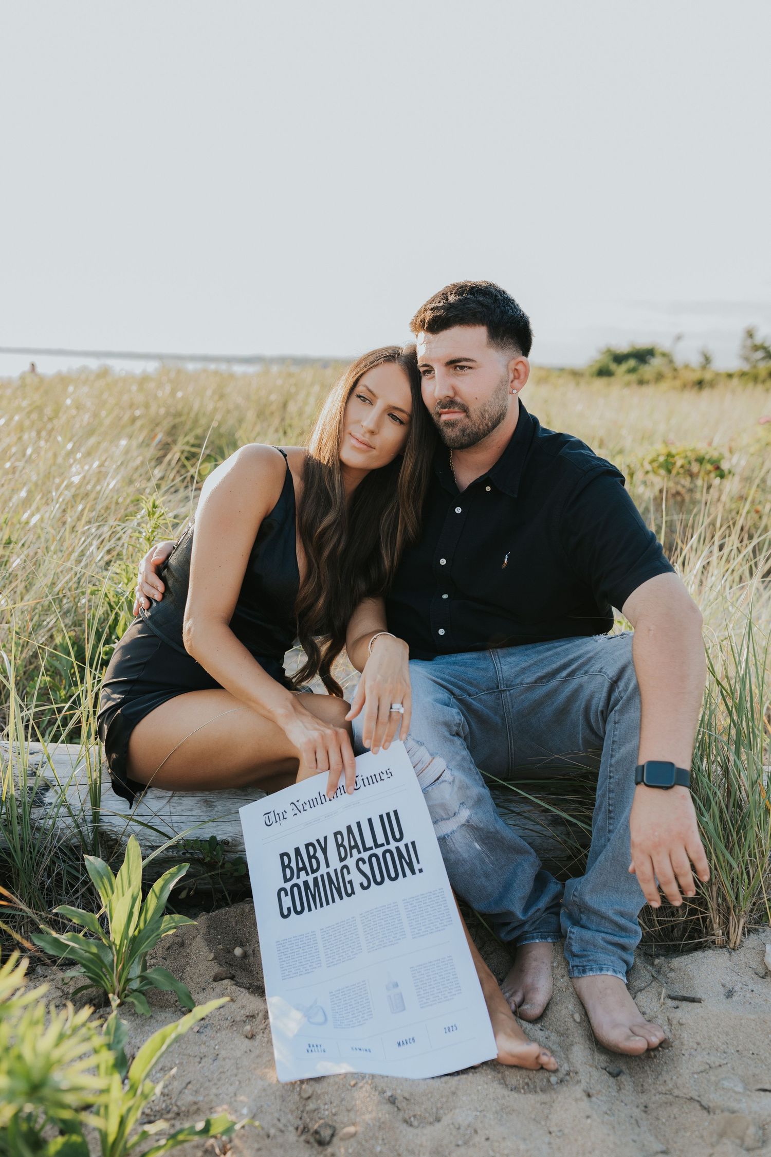 Couple sitting on sandy dunes with Baby Balliu Coming Soon newspaper announcement in front of them.