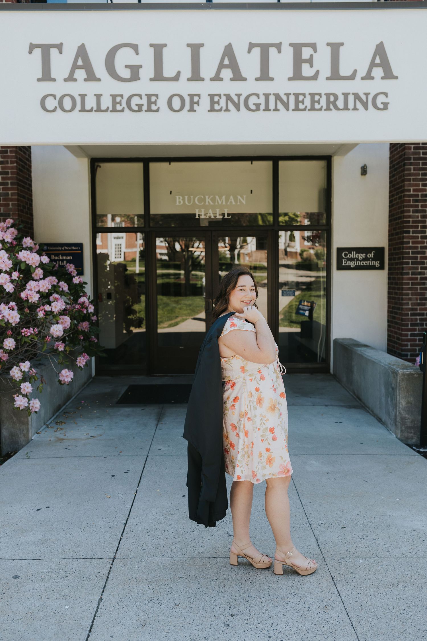 Graduate in floral dress holding gown outside Tagliatela College of Engineering Buckman Hall entrance.