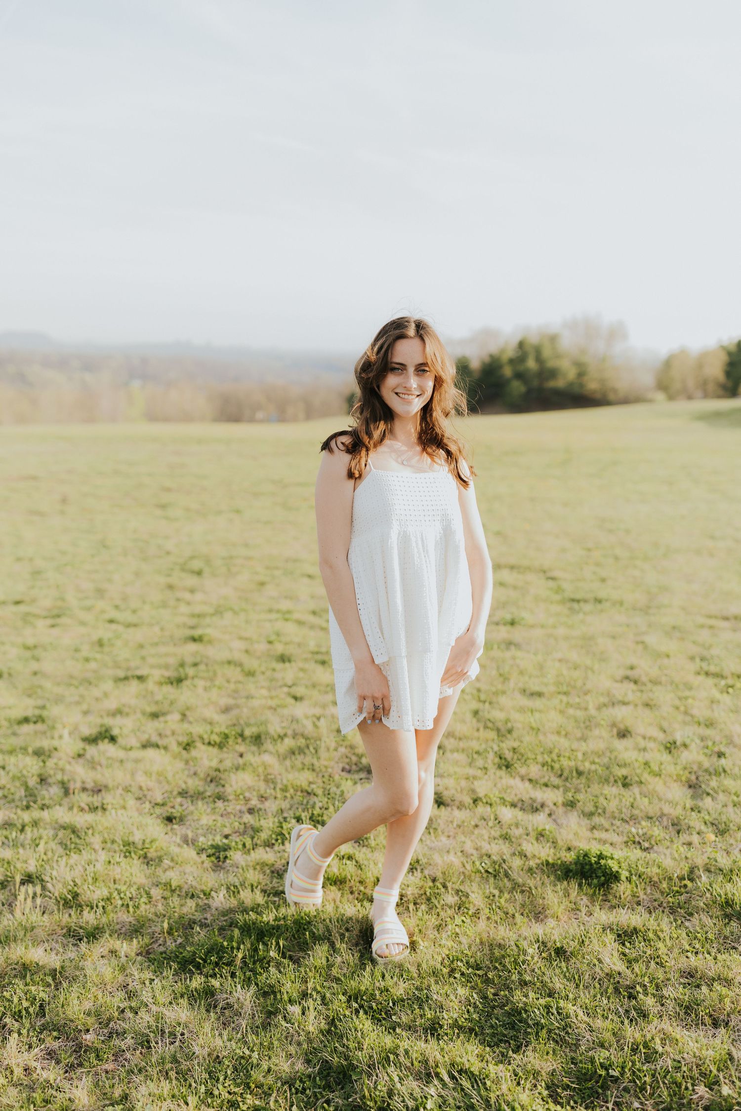 Young woman in white sundress smiling in sunny green field with rolling hills in background.