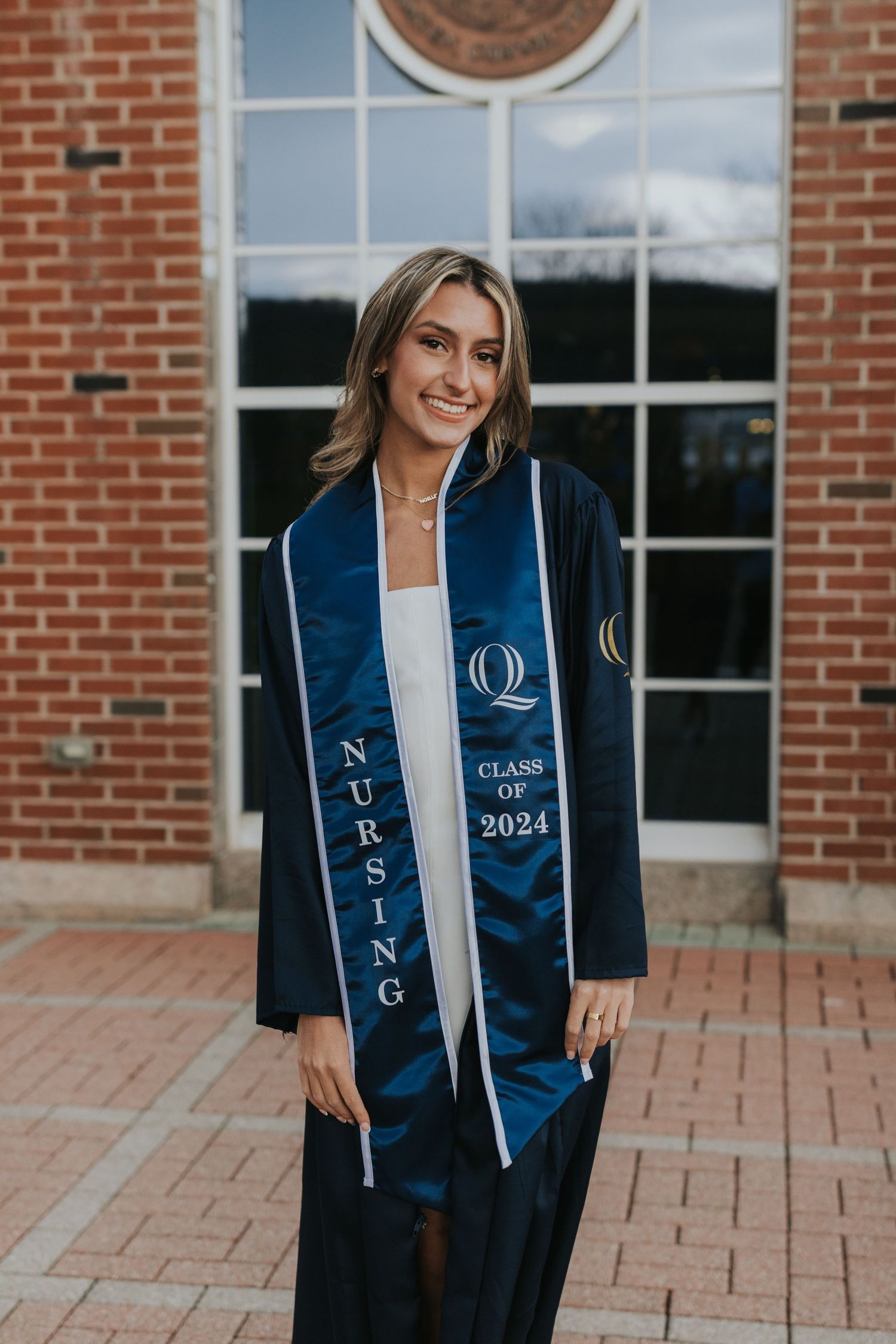 Smiling nursing graduate in navy cap and gown with Class of 2024 stole poses at brick library.
