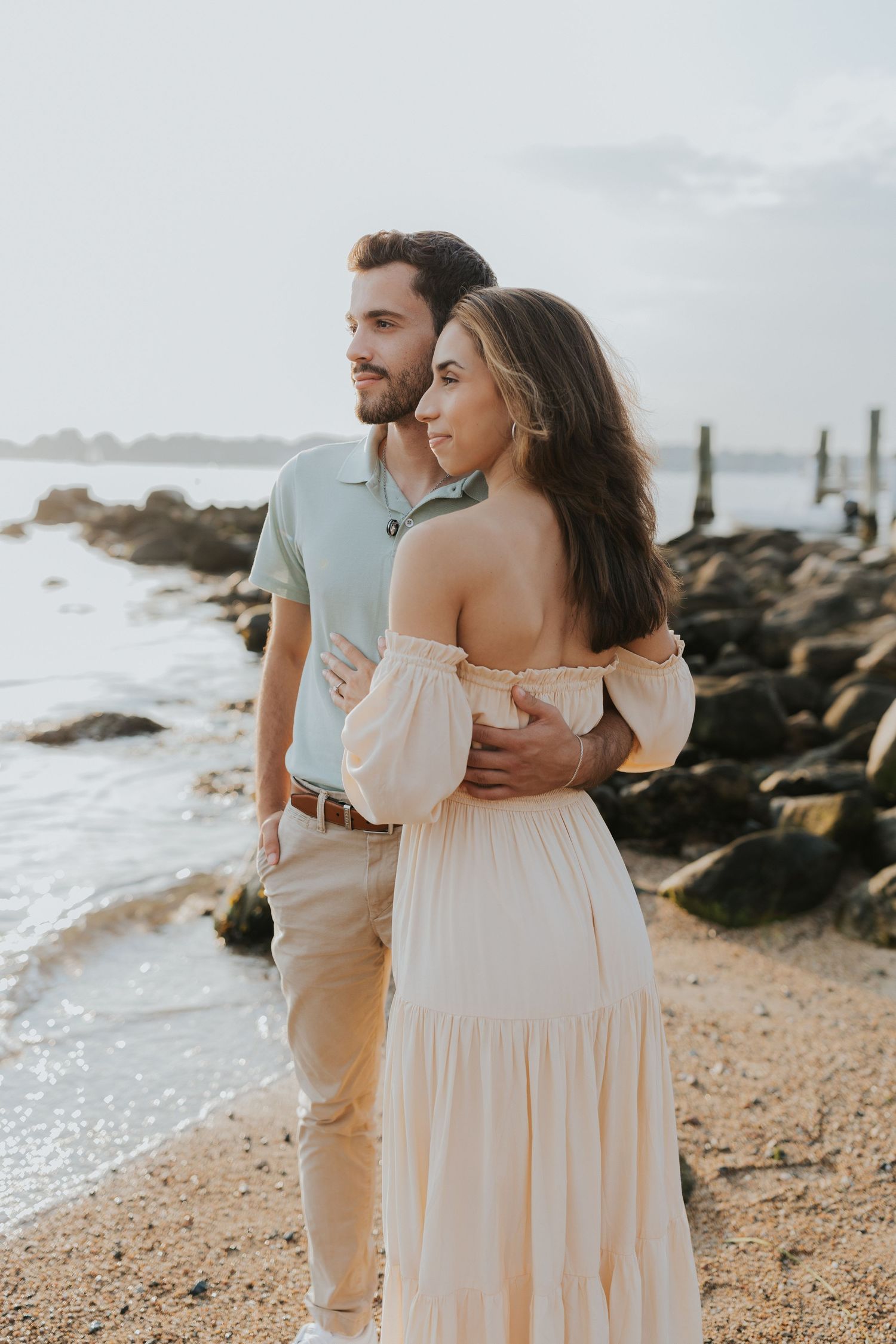 Couple standing together on a sandy beach near mossy rocks, man embracing woman in cream off-shoulder dress.