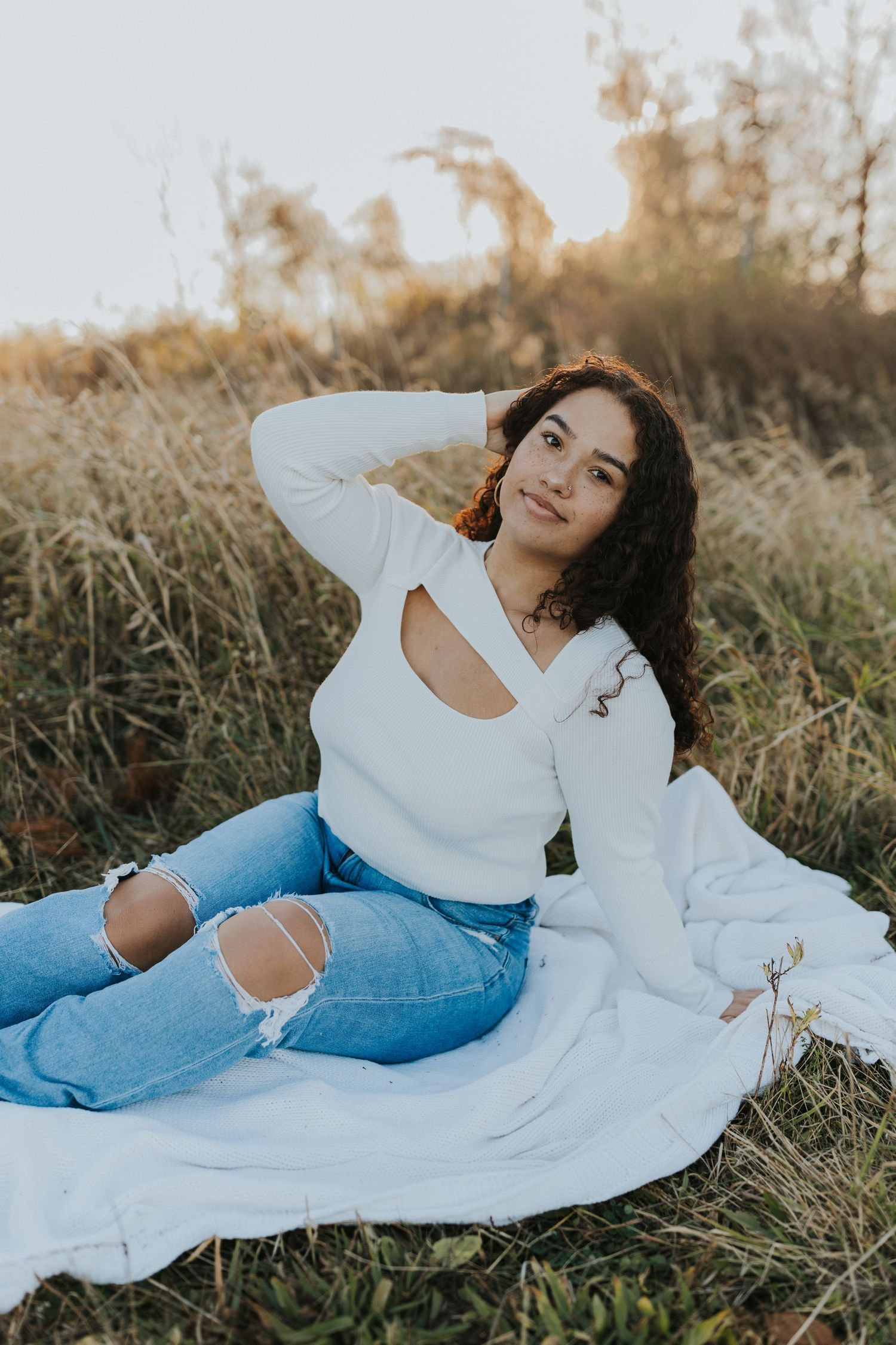 Young woman with curly hair posing on white blanket in field during golden hour sunset.