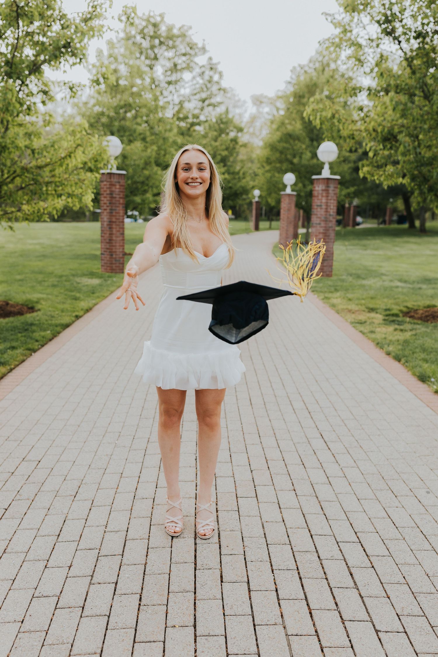 Joyful graduate in white dress tossing mortarboard cap on a brick tree-lined campus path.