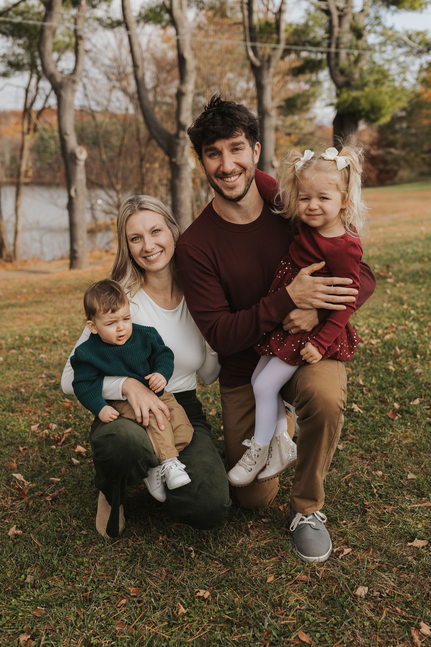 Happy family of four posing together on grass with autumn trees and lake in background.