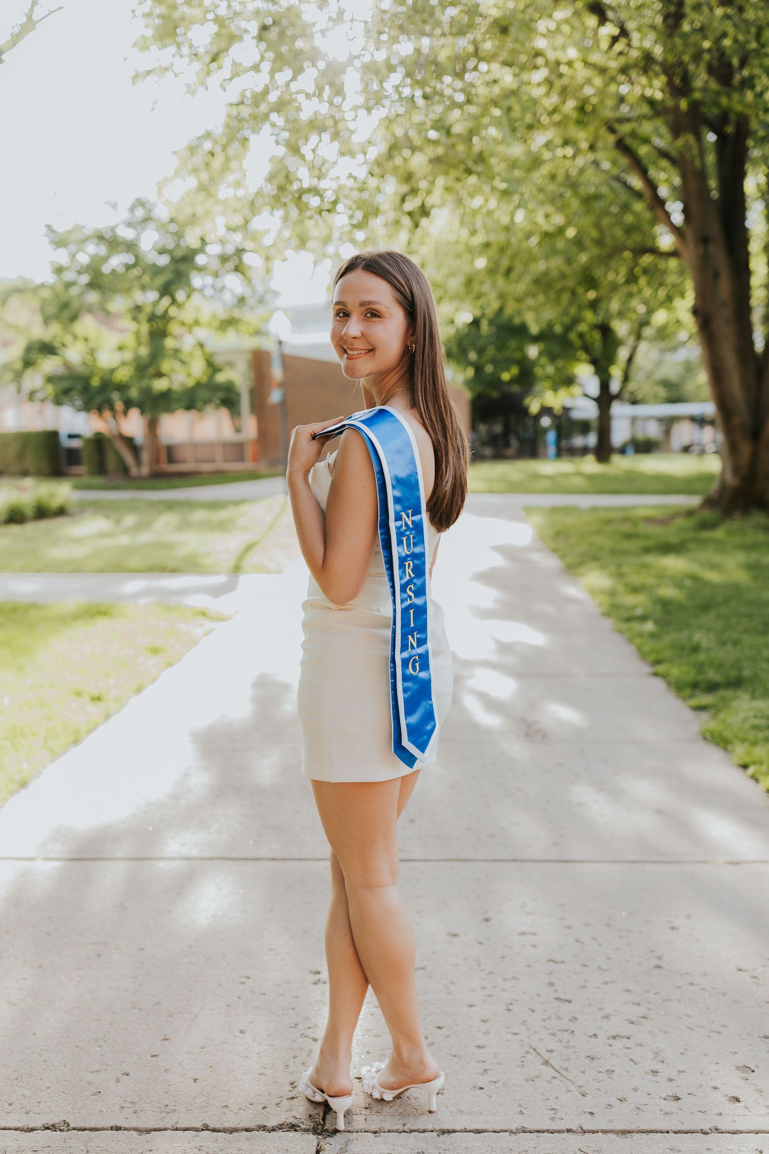 Smiling nursing graduate in white dress poses with blue sash on campus path.