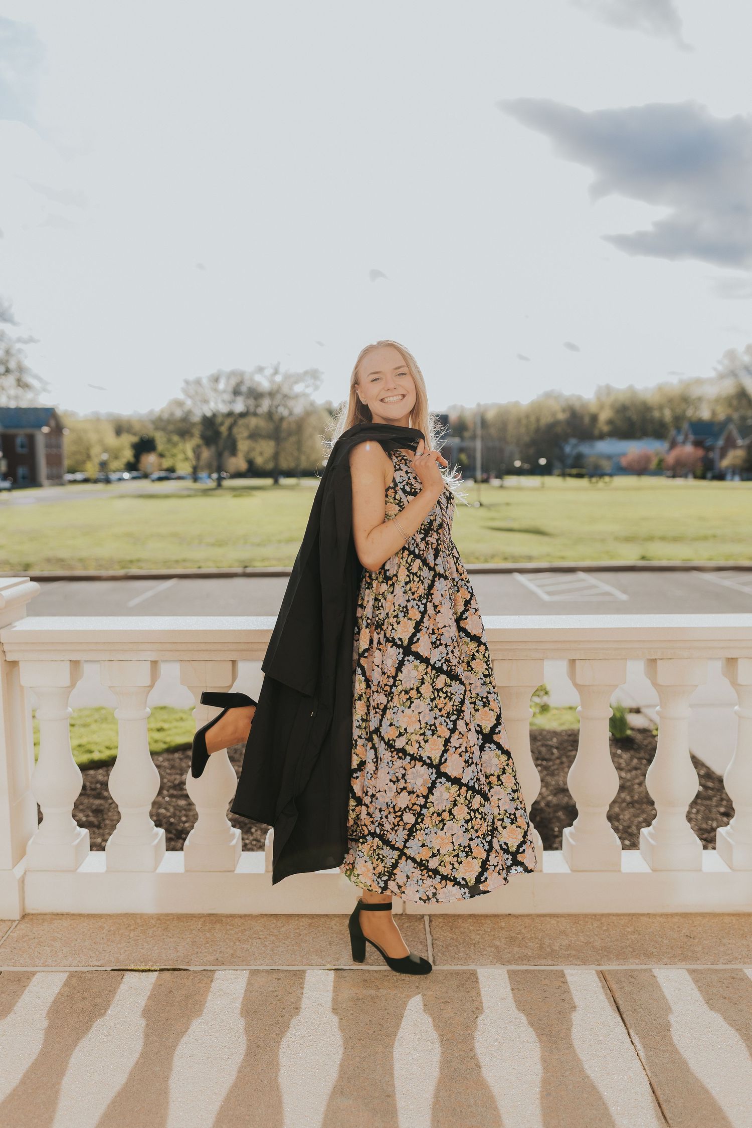 Smiling blonde woman in floral midi dress holding black blazer poses on white balustrade outdoors.