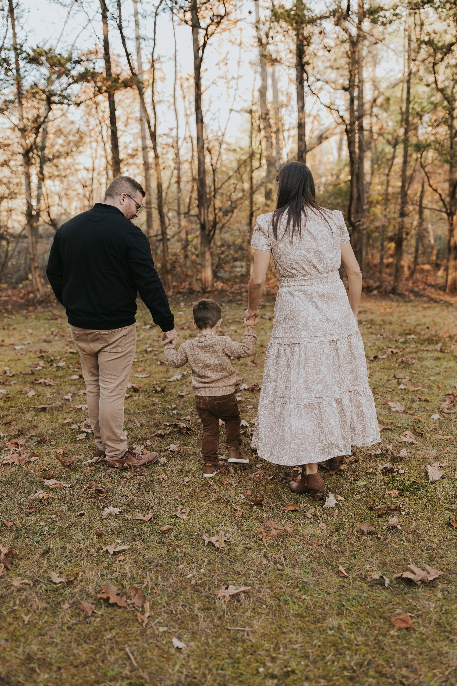 A young family of three walks hand-in-hand through a colorful autumn forest during a fall photo session.
