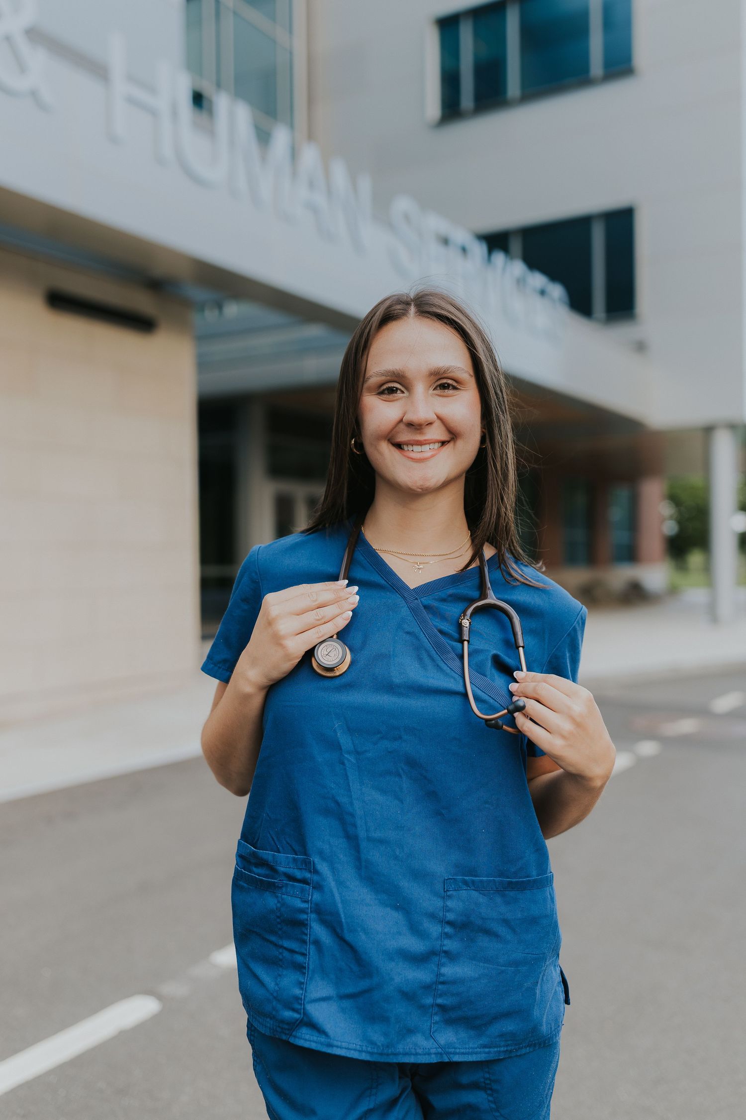 Happy female nurse in blue scrubs with stethoscope posing outside Human Services building.