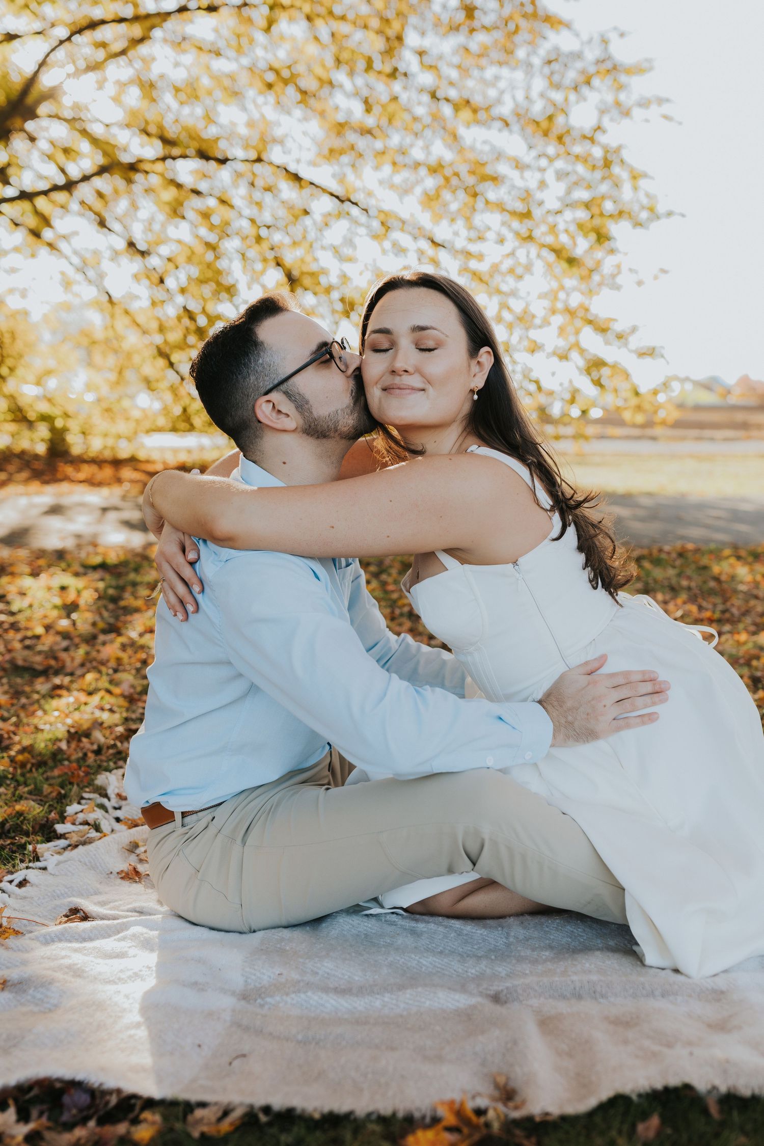 Couple sharing a kiss on a blanket under golden autumn trees in a park setting.