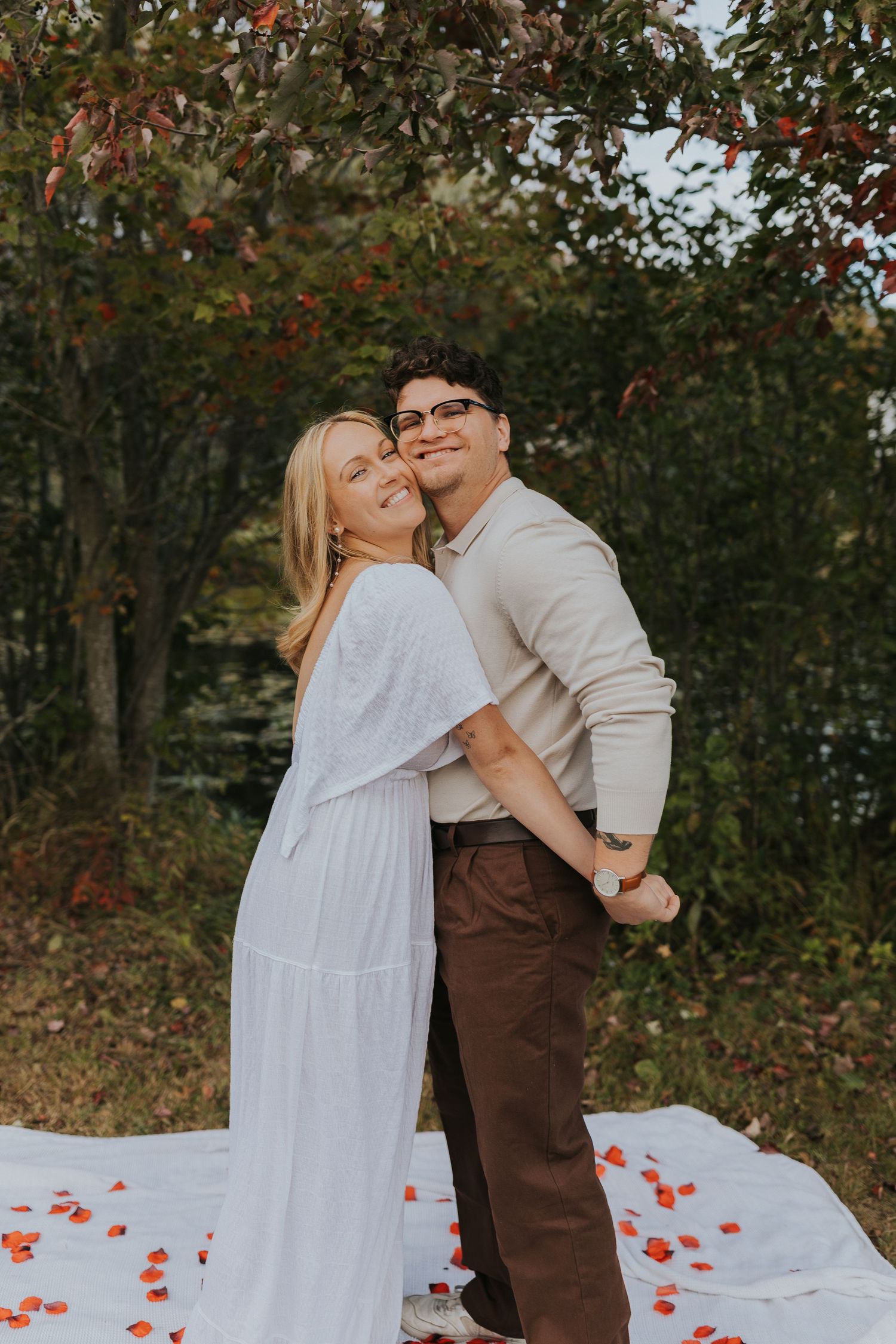 Smiling engaged couple posing under autumn foliage near a pond with rose petals on blanket.