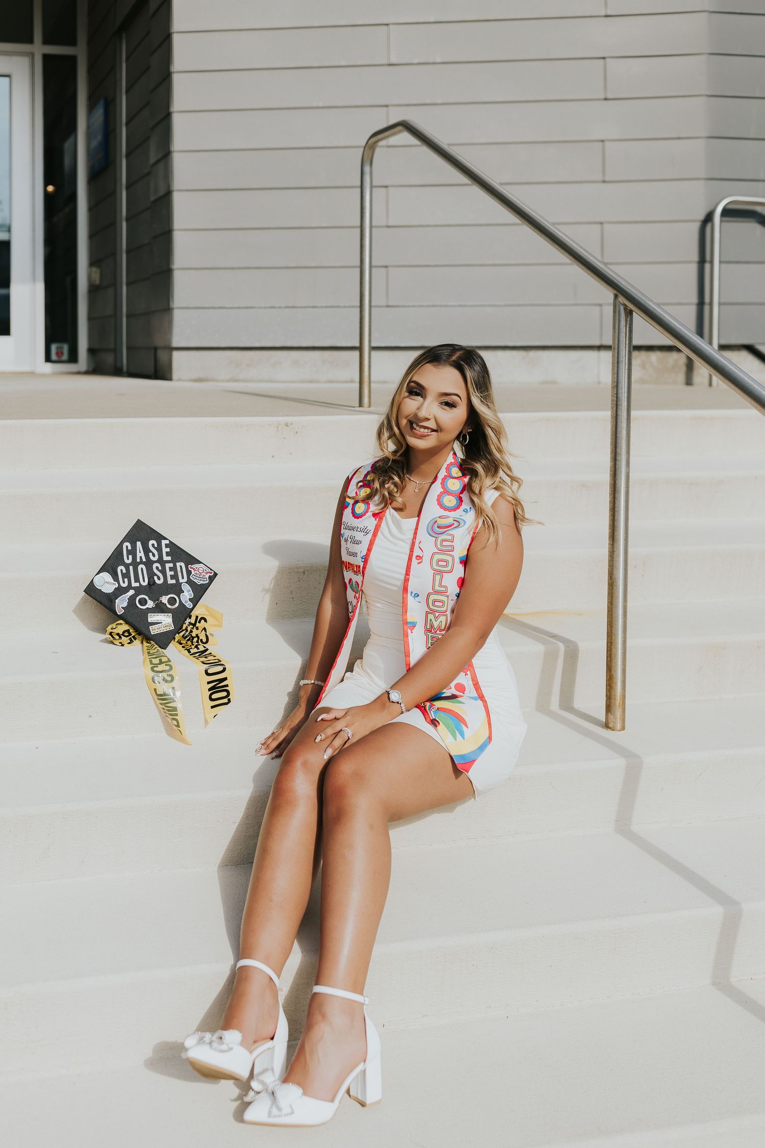 Smiling graduate in white dress and colorful stole sits on white steps with Case Closed forensic graduation cap.