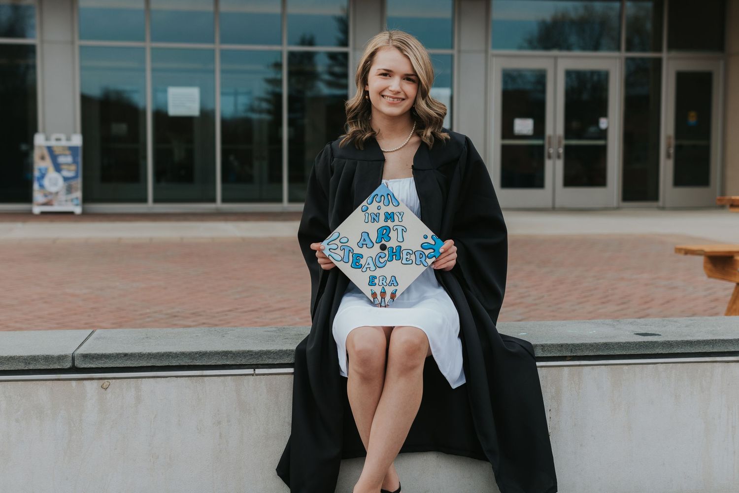 Smiling female graduate sits outdoors holding decorated graduation cap without gown.