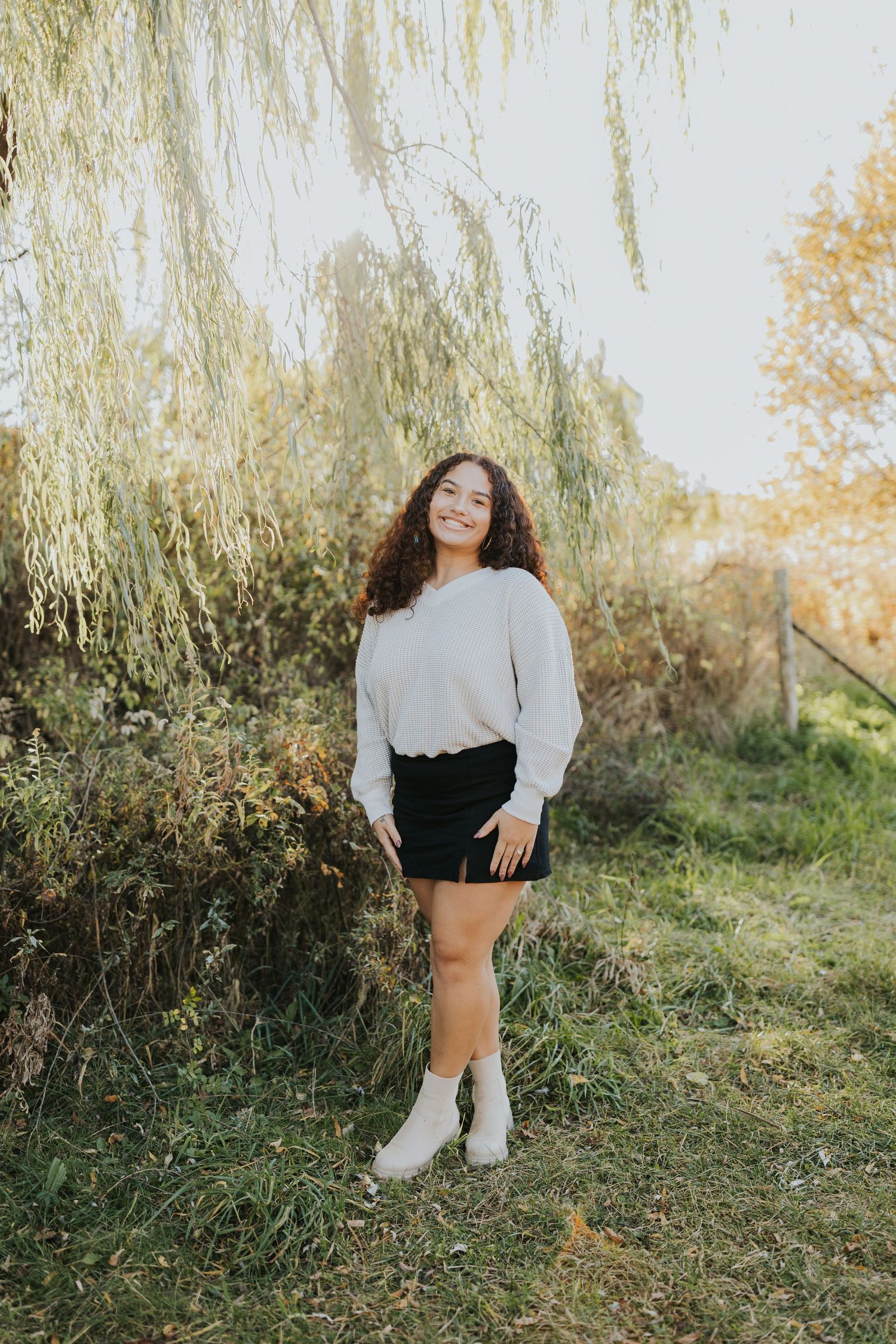 Young woman with curly hair in cream sweater and black skirt smiling under willow tree.