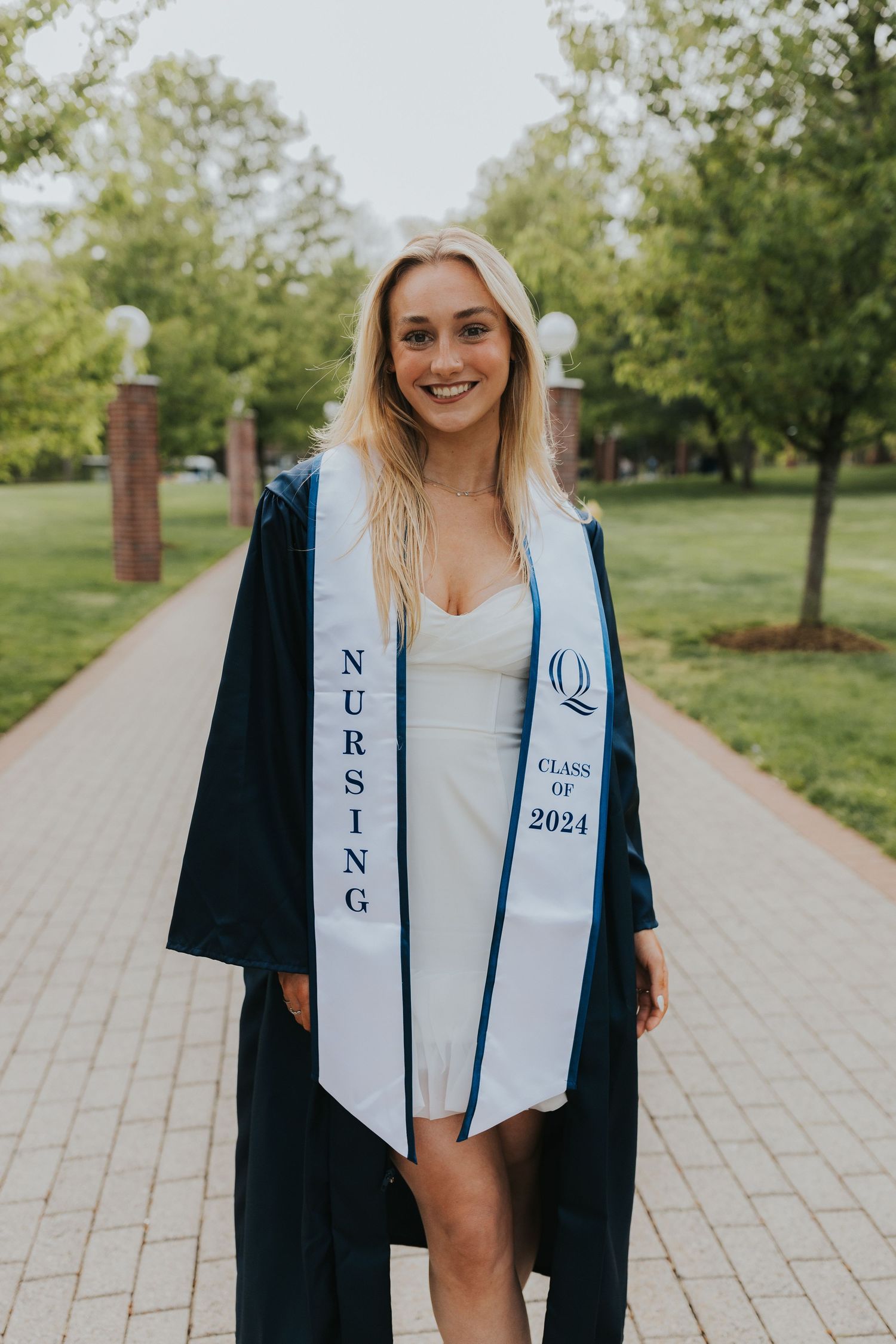 Blonde graduate smiles in navy gown with Nursing Class of 2024 stole on campus path.