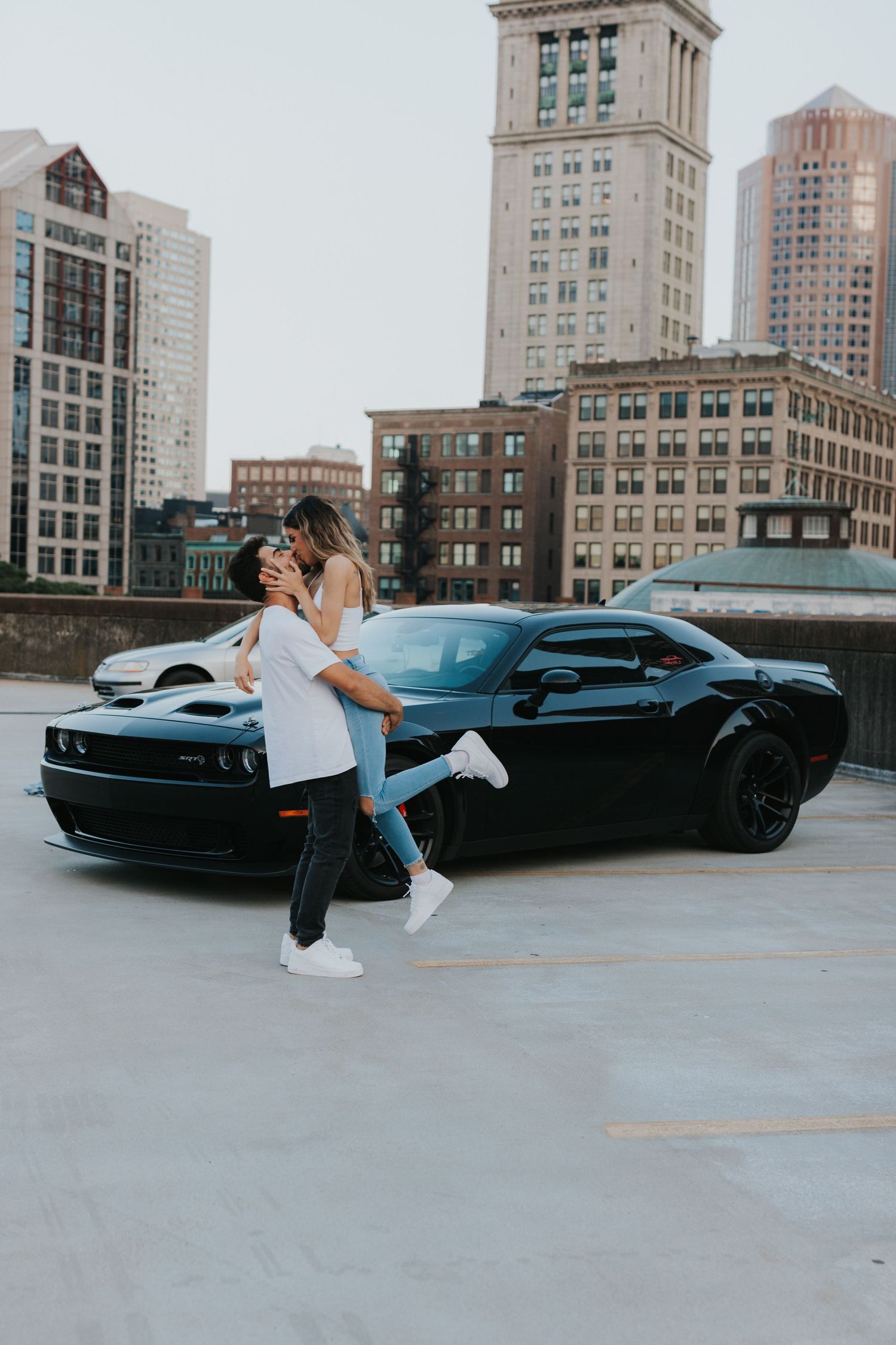 Man lifting woman in a romantic embrace in front of a black Dodge Challenger on a city rooftop.