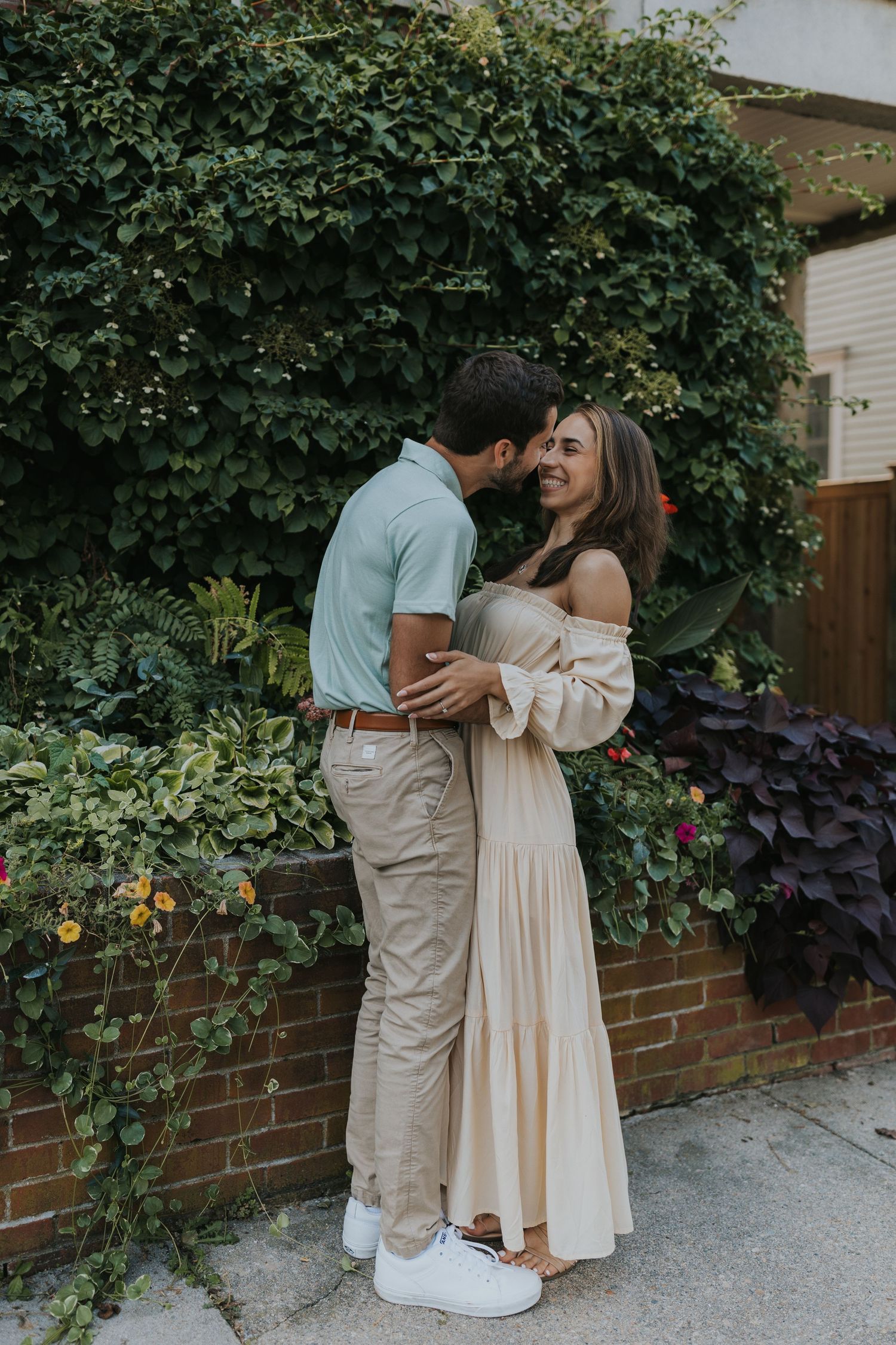Romantic couple nearly kissing against lush green ivy wall, man in blue polo embracing woman in flowy cream dress.