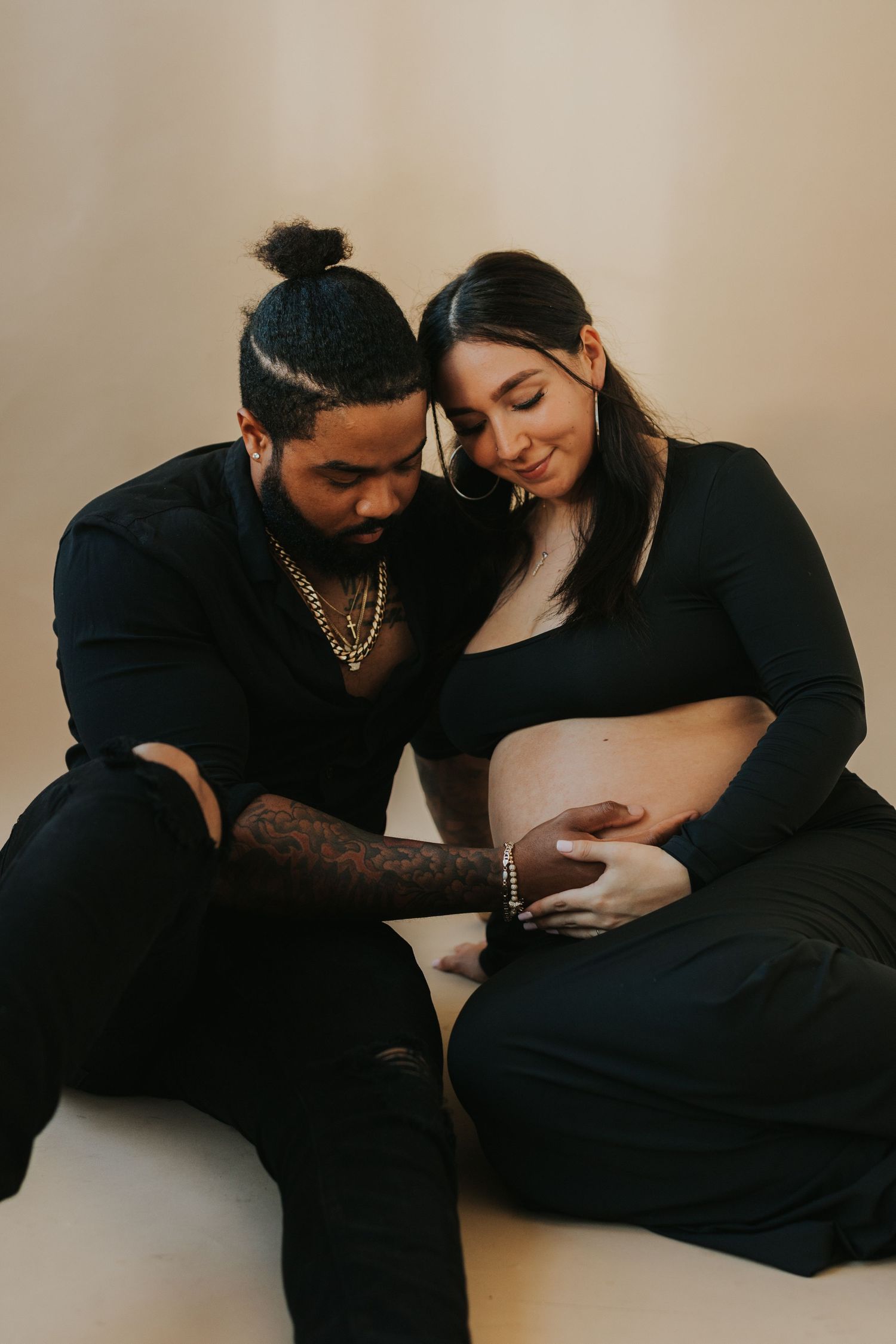Couple sitting on floor in black outfits, gently touching pregnant woman's bare belly.