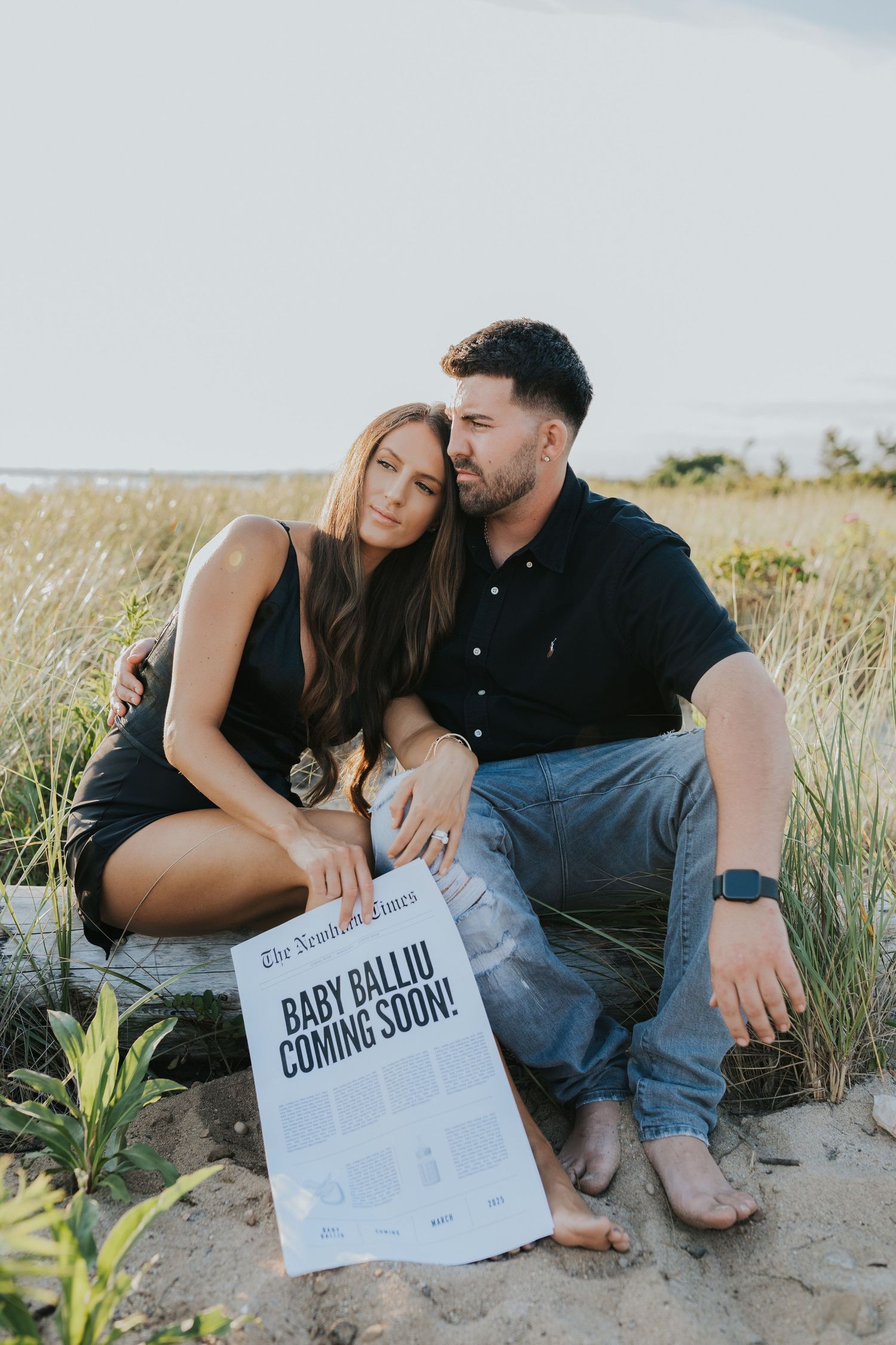 Couple sitting together on sandy beach dunes with Baby Balliu Coming Soon newspaper announcement.