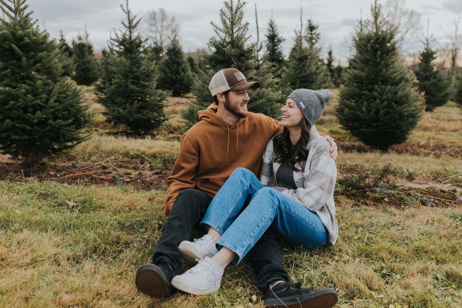 Couple sitting together on grass at a Christmas tree farm, smiling at each other.