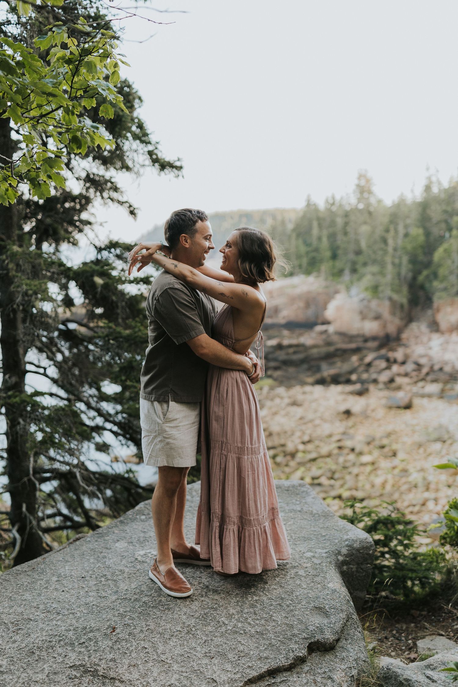 Couple embracing on a rocky overlook surrounded by pine trees and coastal scenery.