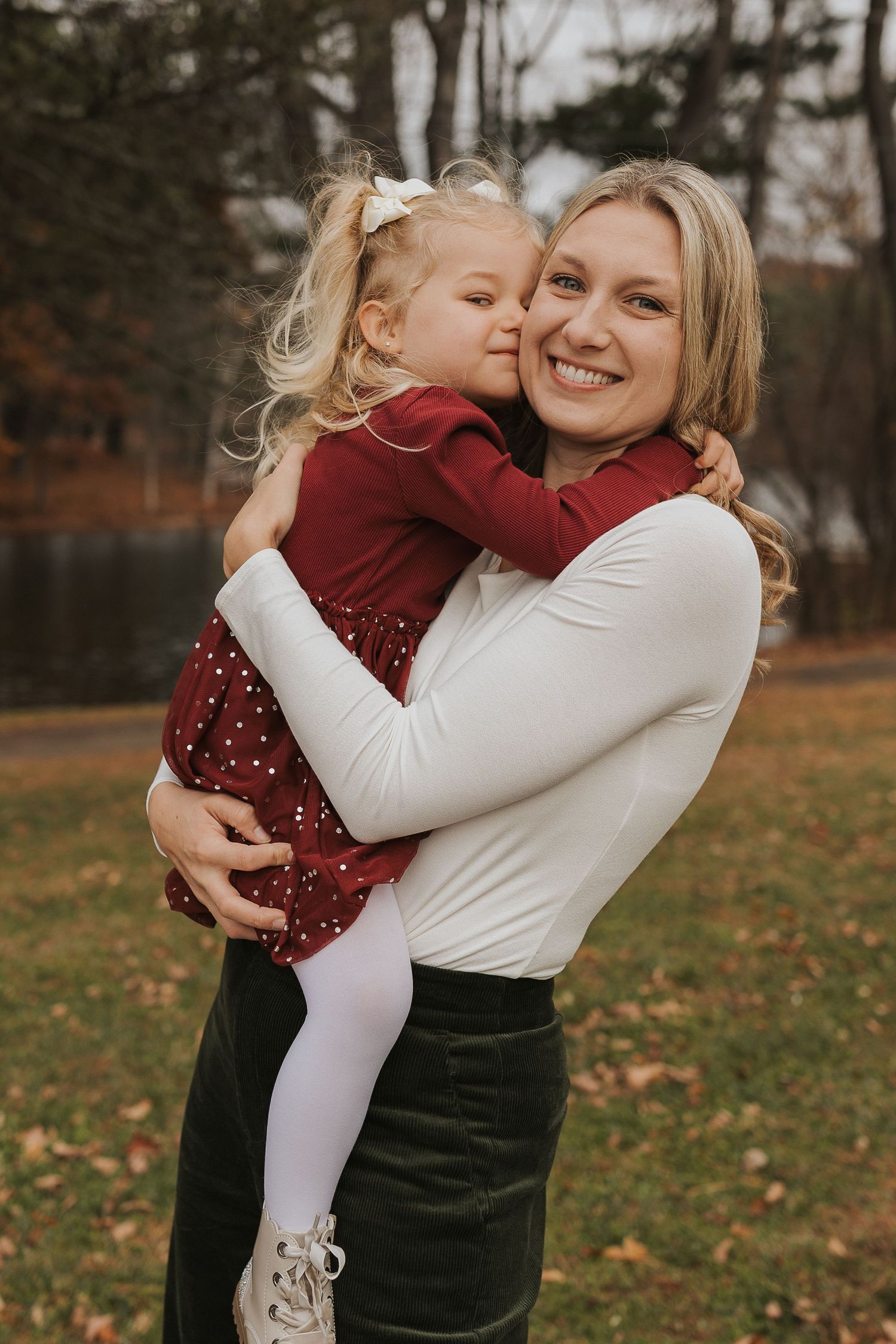 Blonde mother smiling as her young daughter in a red dress gives her a kiss outdoors.
