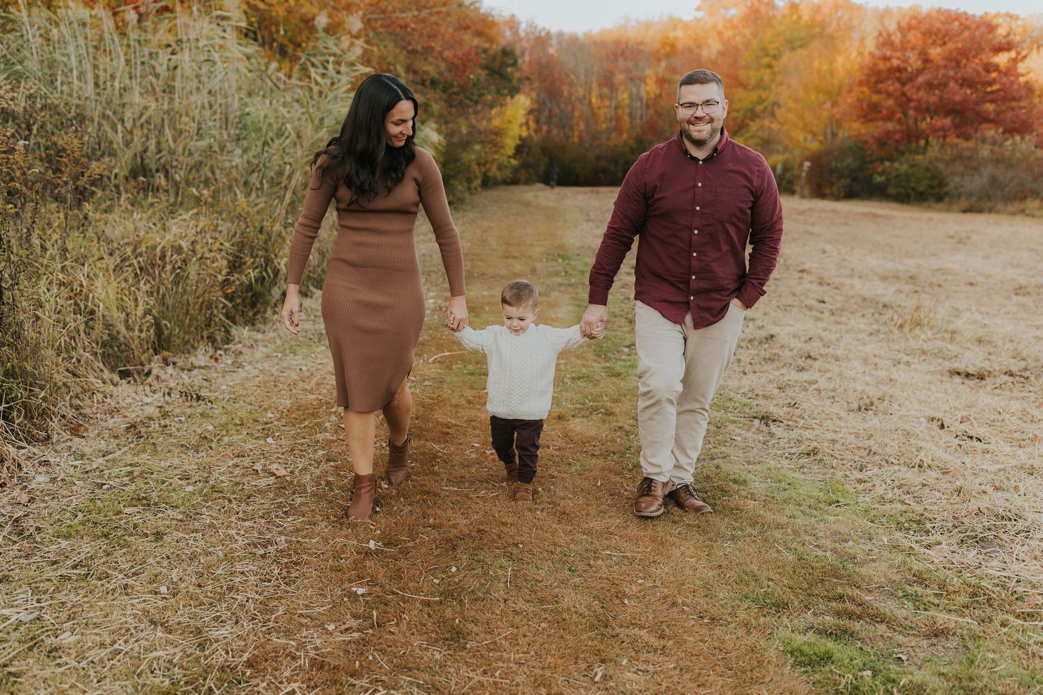 Family of three walking hand in hand through golden autumn field during fall family photo session.