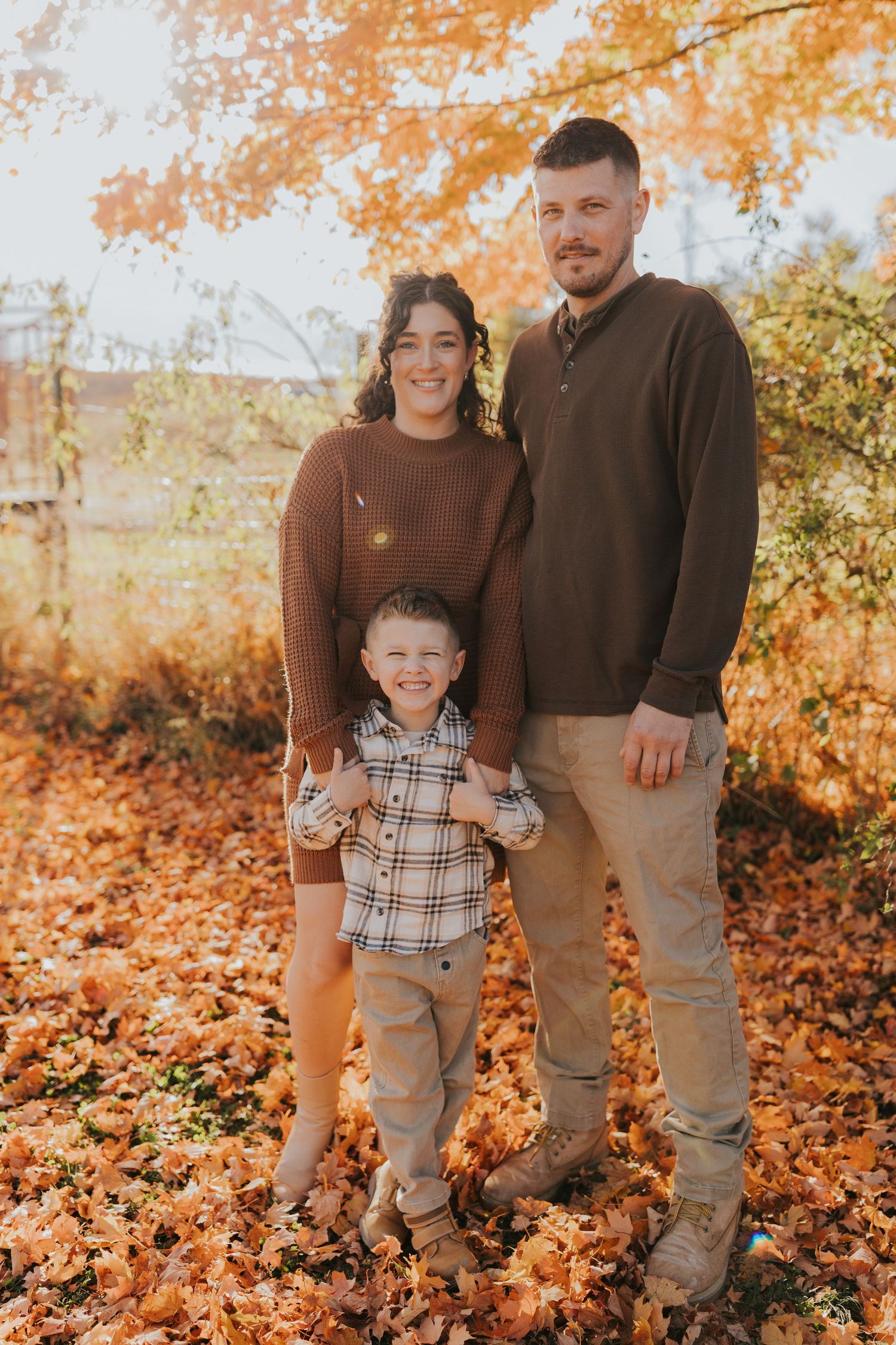 A family of three stands together smiling among bright orange autumn leaves in a wooded area.