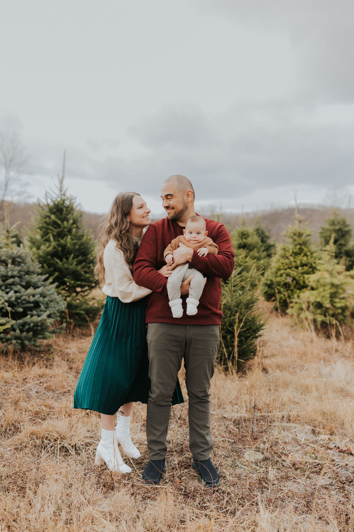 Family of three standing among Christmas trees, father holding infant while parents gaze at each other.