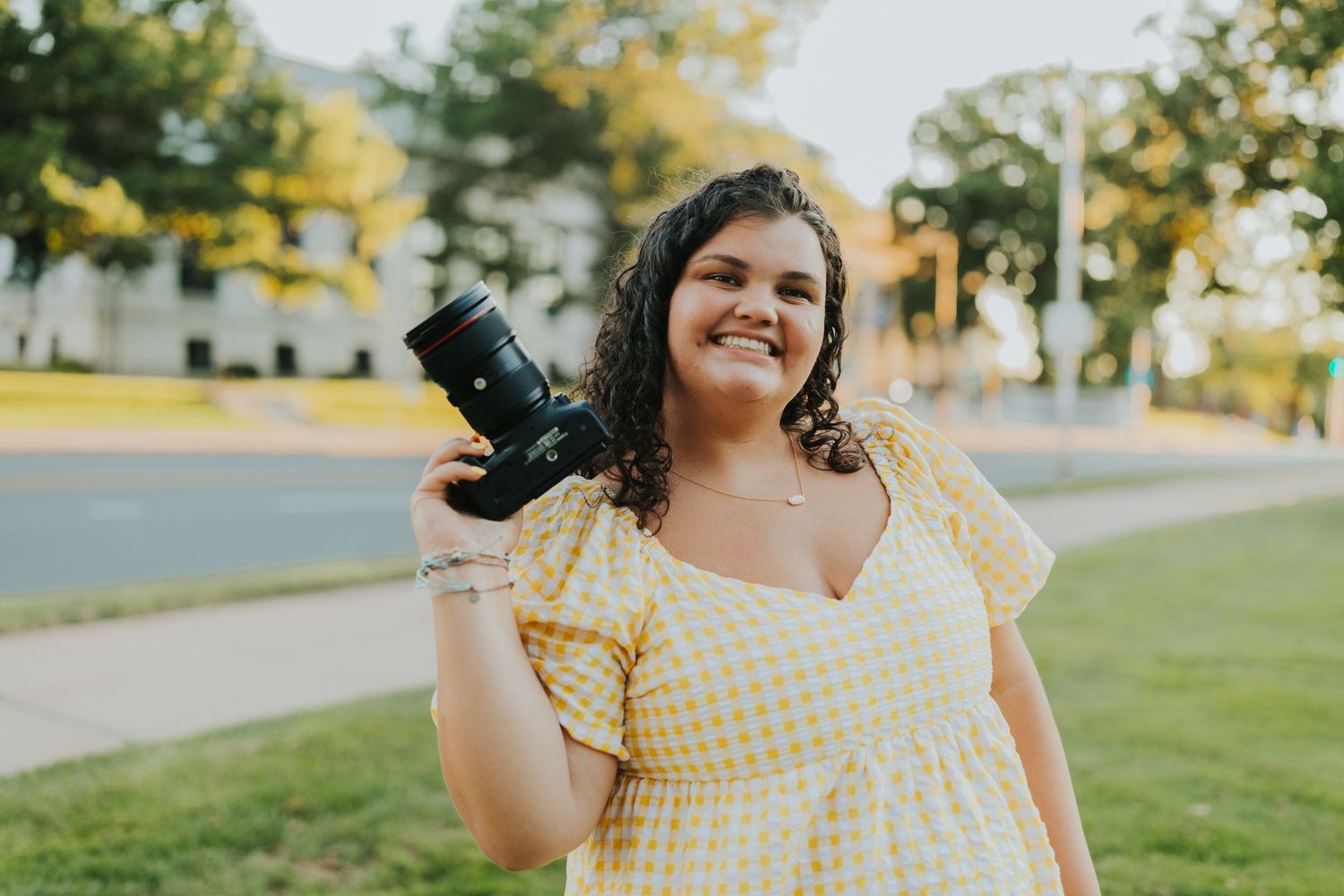 Happy photographer in yellow gingham dress holding camera aloft on a sunny green outdoor setting.
