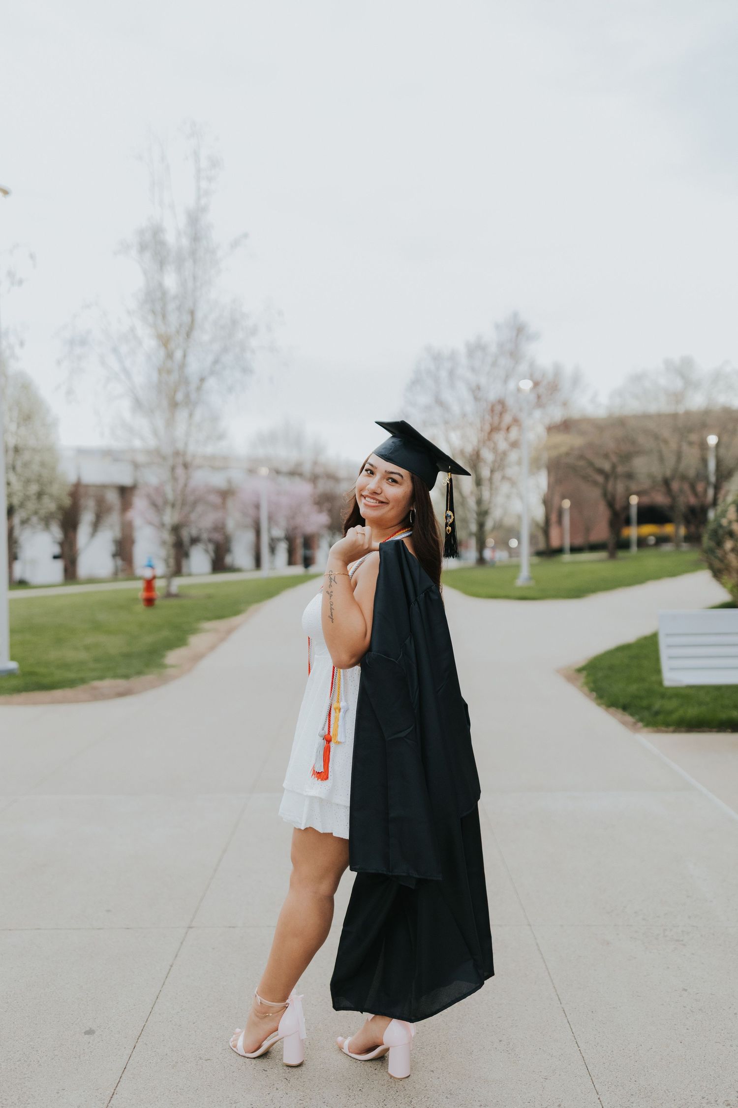 Graduate poses in black cap and gown on campus walkway, smiling over shoulder.