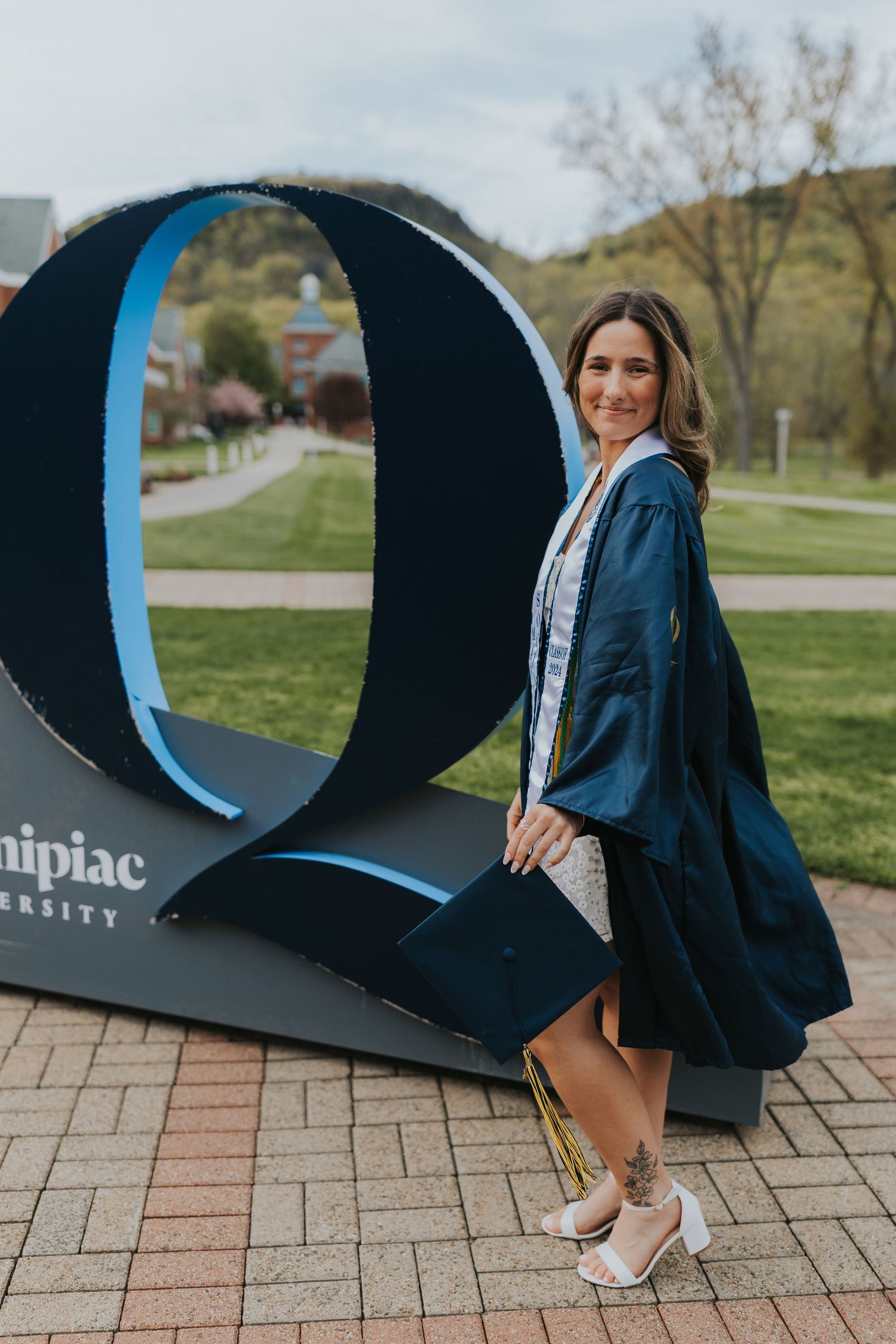 Graduate poses in navy cap and gown beside Quinnipiac University large Q letter sign on campus.