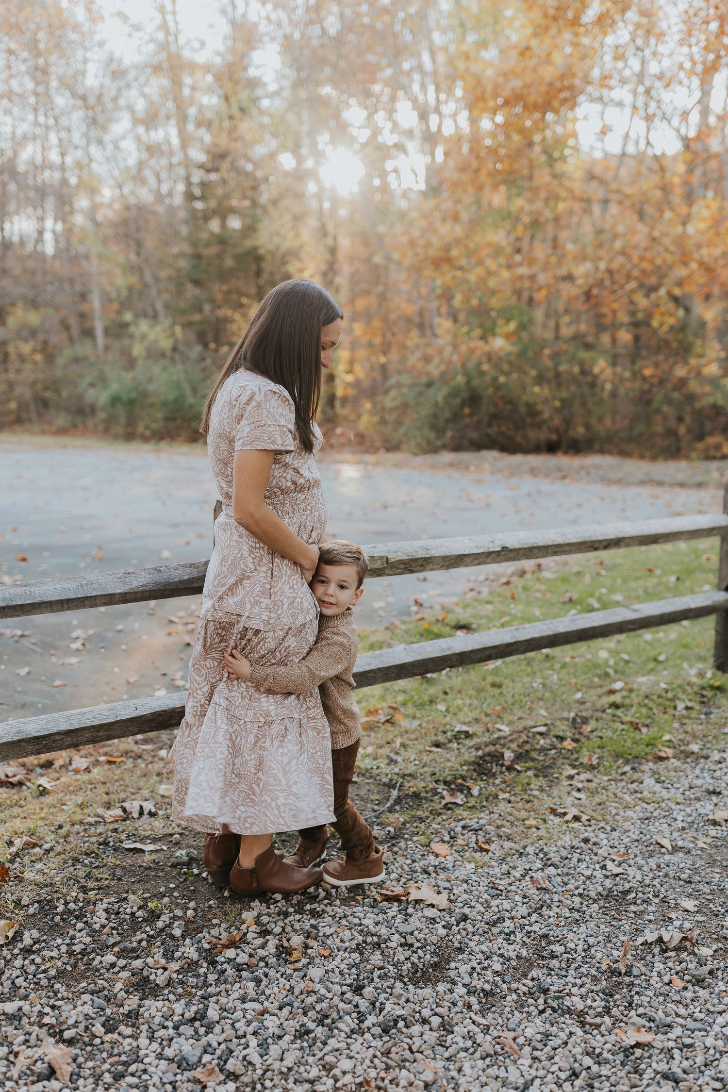 Pregnant mother in floral dress hugging young child near wooden fence in fall foliage setting.