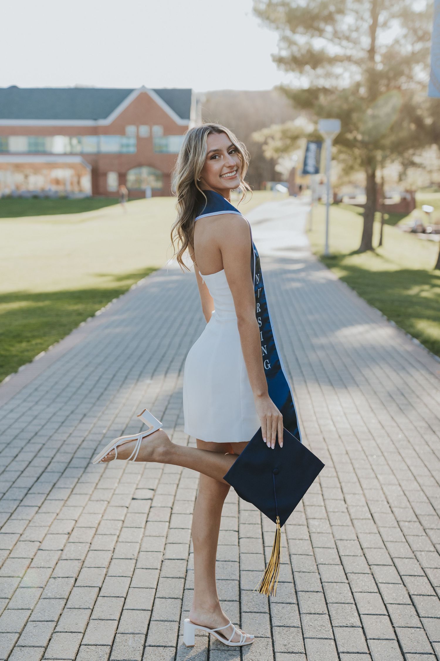 Smiling graduate in white dress holding cap with blue stole on sunlit brick campus pathway.