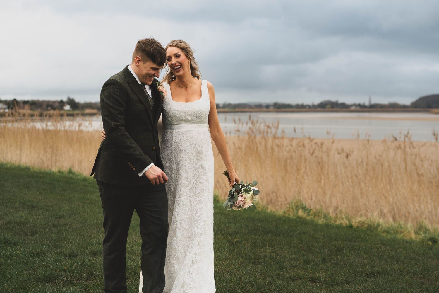a bride and groom walk along a riverside in the cotswolds