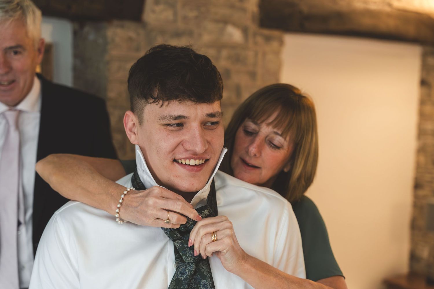 the mother of the bride helps a groom put on his tie at a wedding in the cotswolds