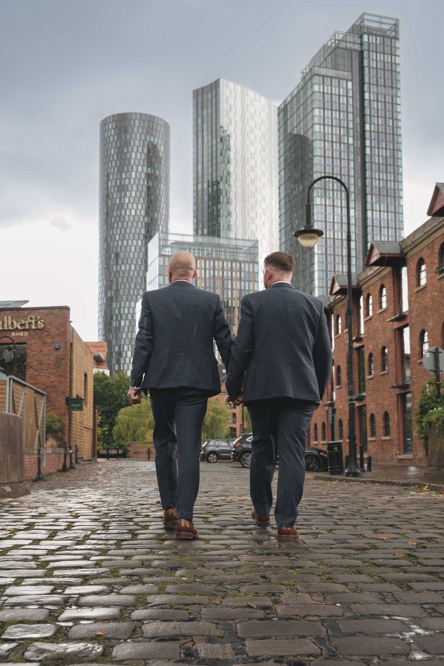 2 grooms walk towards large buildings in the city of Manchester