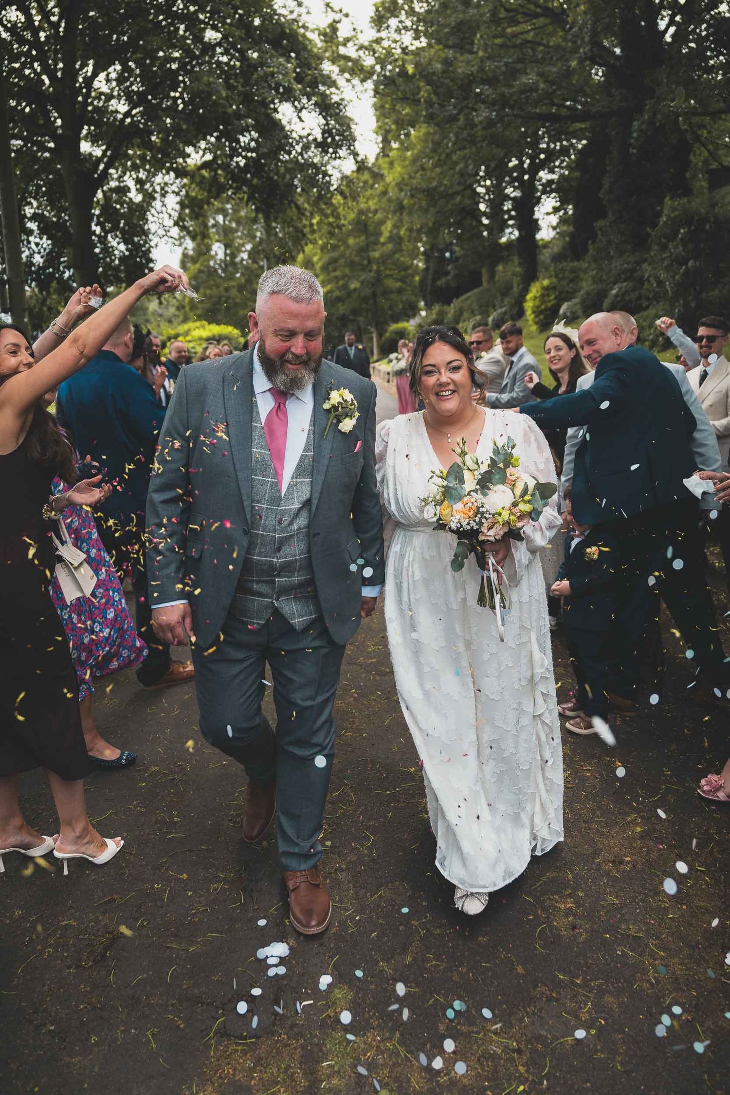 a bride and groom smile as they walk through confetti in Stoke