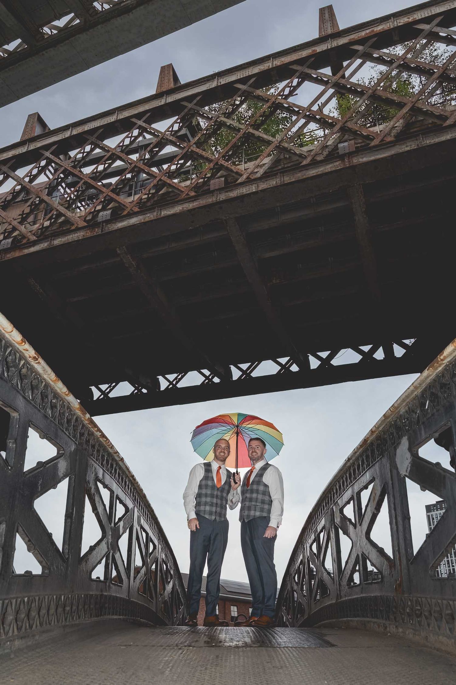 2 grooms stand holding rainbow umbrellas underneath the railway bridges in Manchester