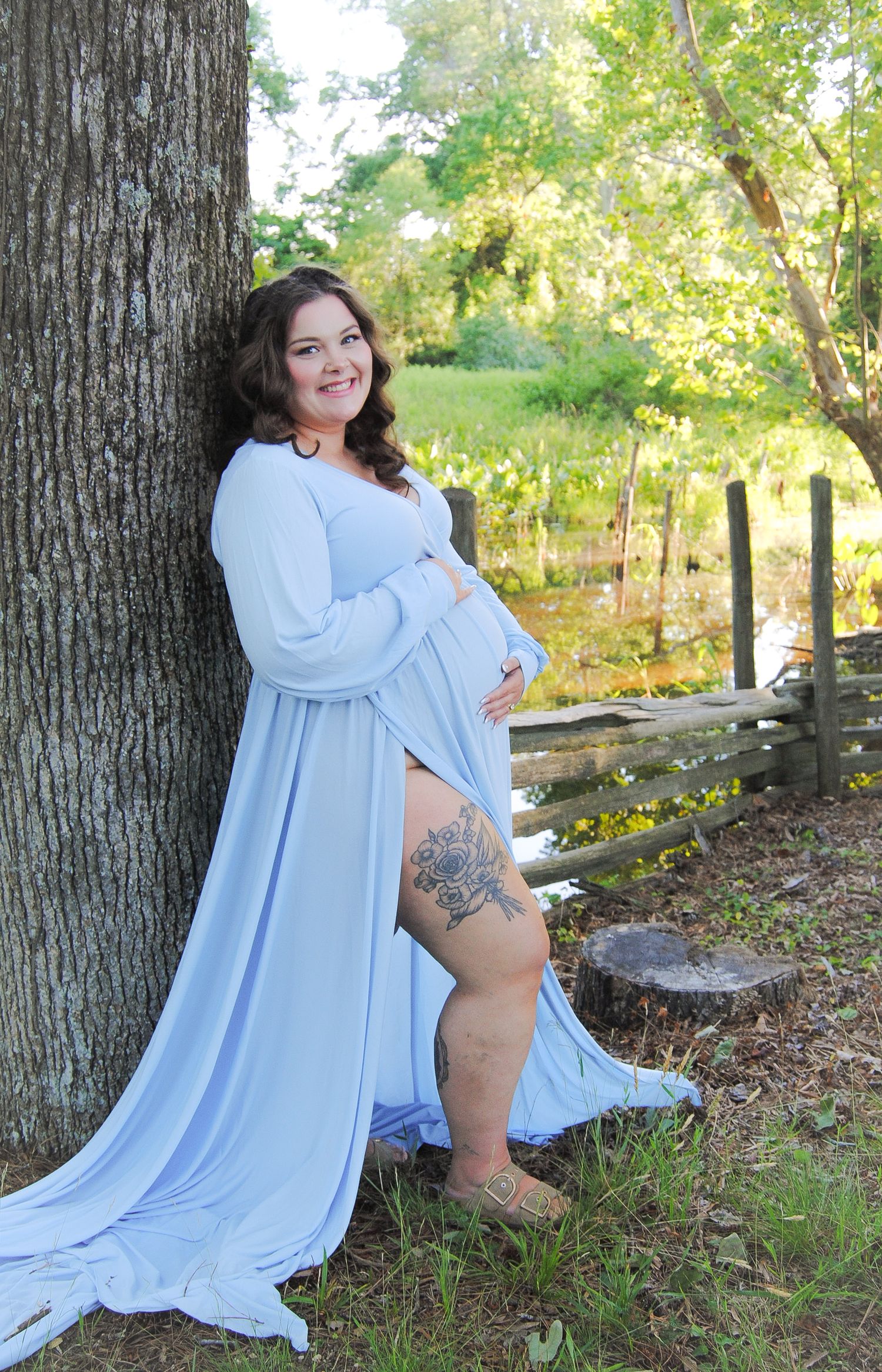 Smiling pregnant woman in light blue maternity gown leaning against tree outdoors with wooden fence and greenery.