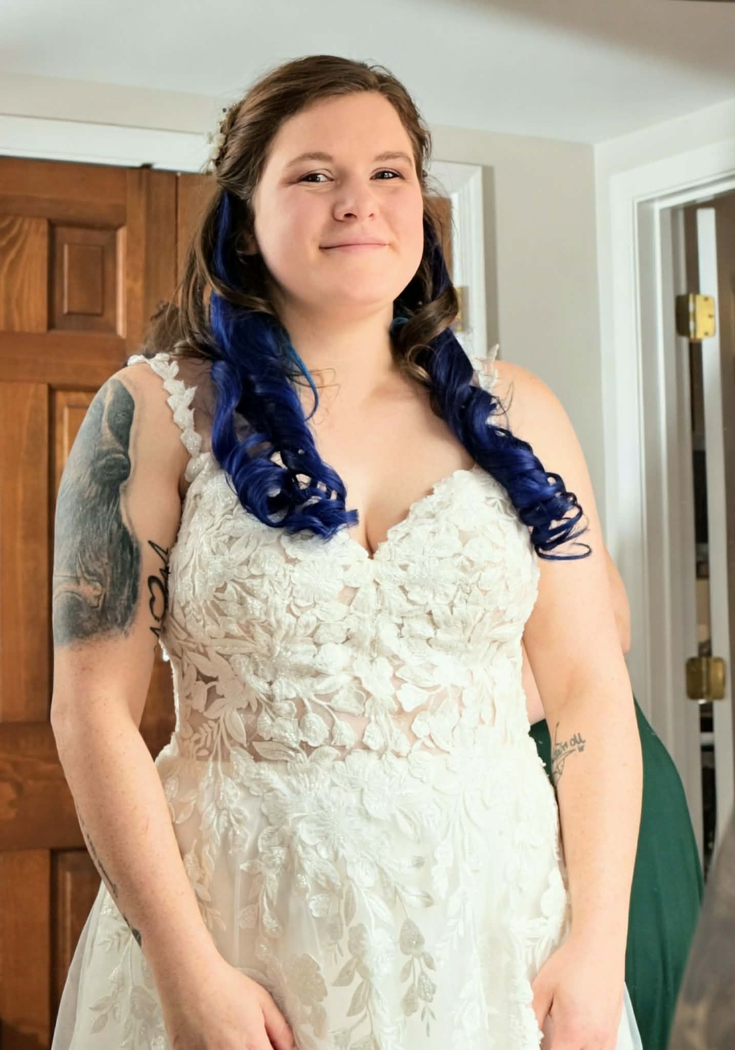 Bride in lace wedding gown with blue highlighted hair and tattoos smiling indoors before ceremony.