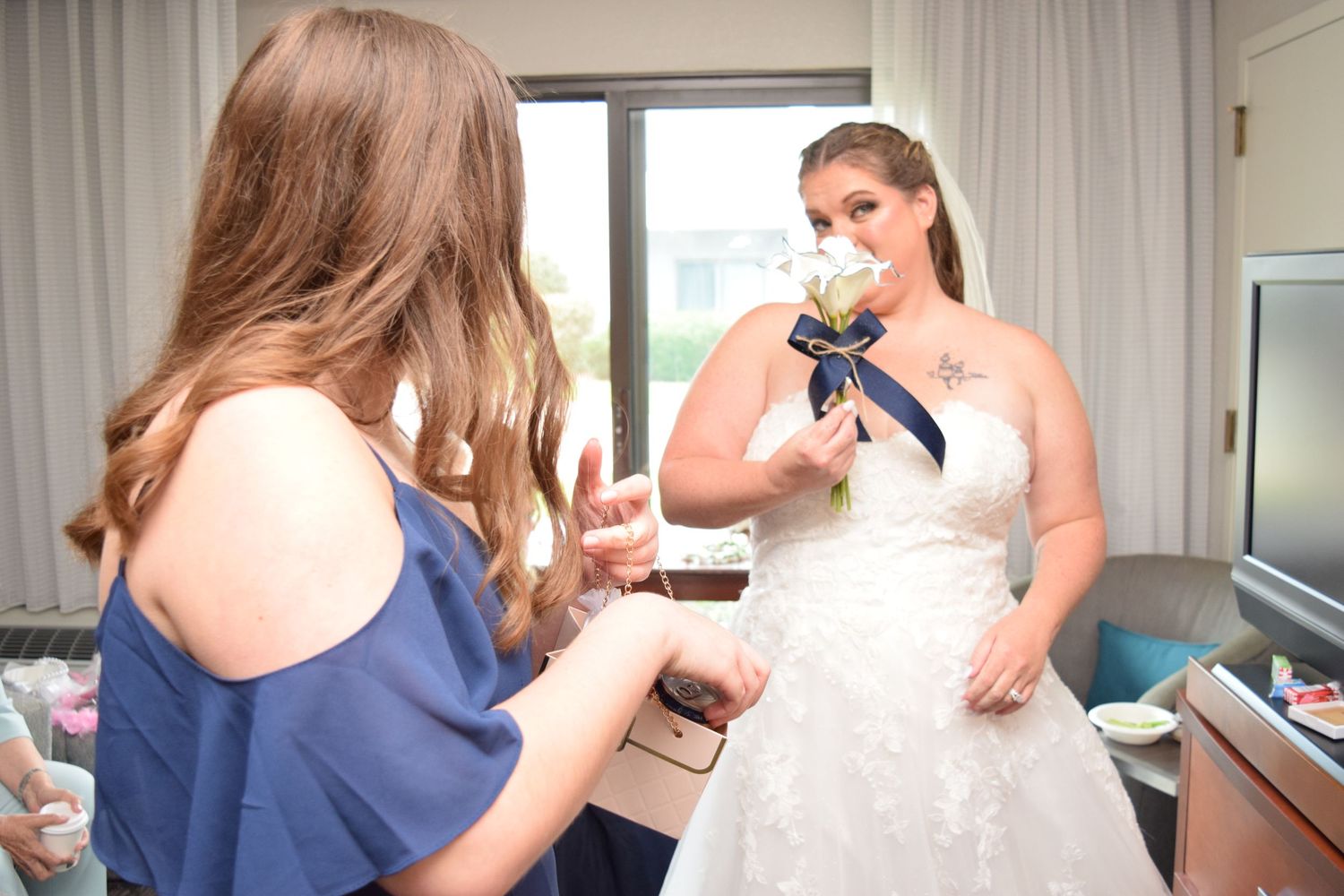 Bride in strapless white gown holding floral boutonniere while chatting with bridesmaid in navy blue dress.