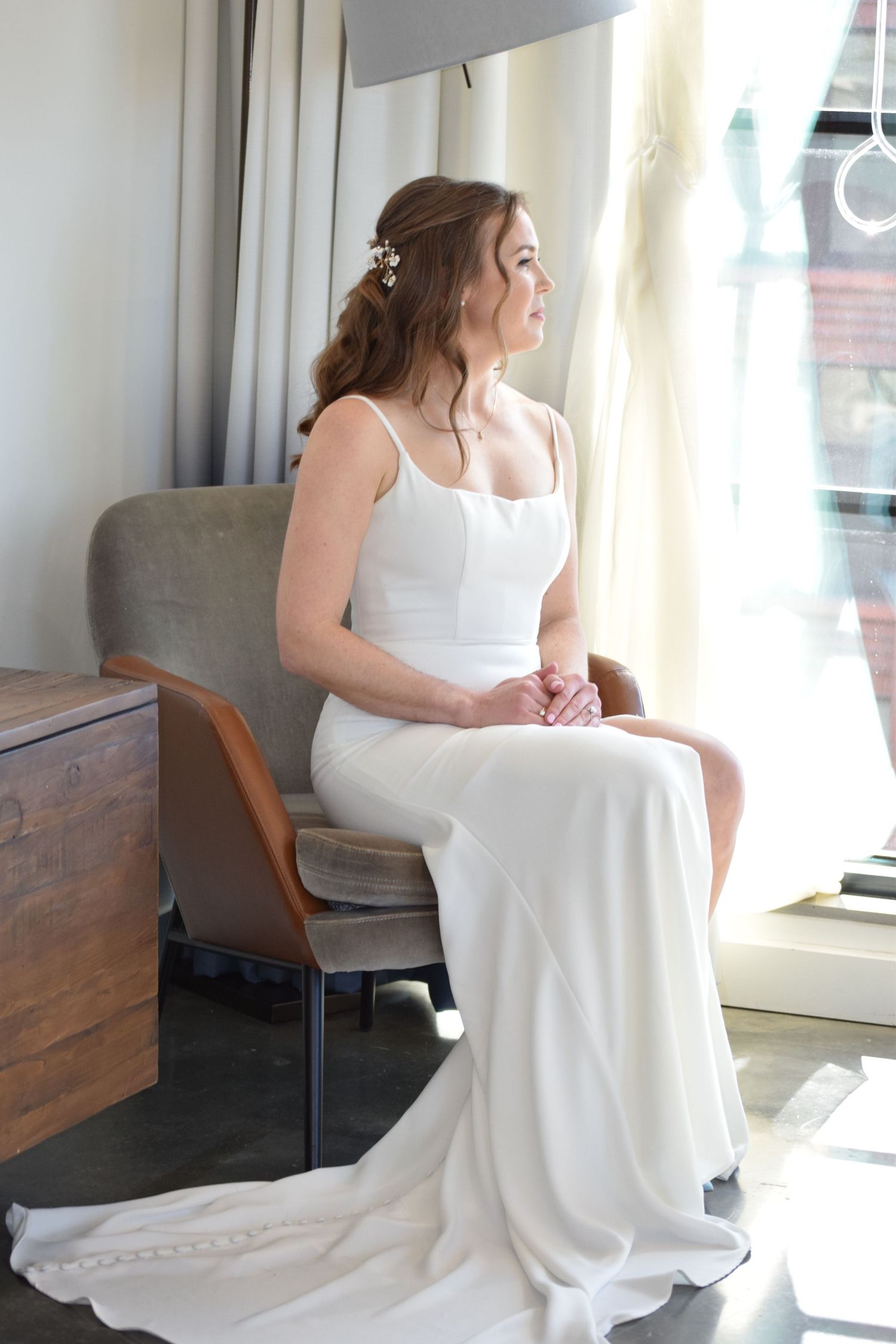 Bride in sleek white wedding dress gazing out window in bright bridal suite.