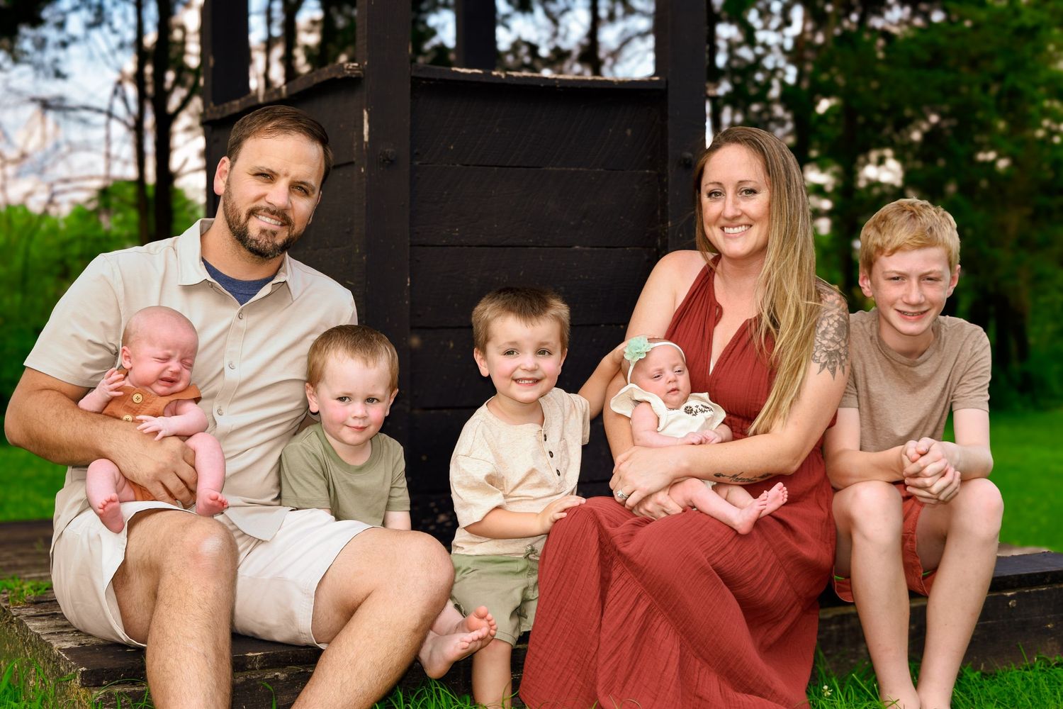 Happy family portrait with parents holding newborn twins and three children sitting outdoors on wooden steps in a park.