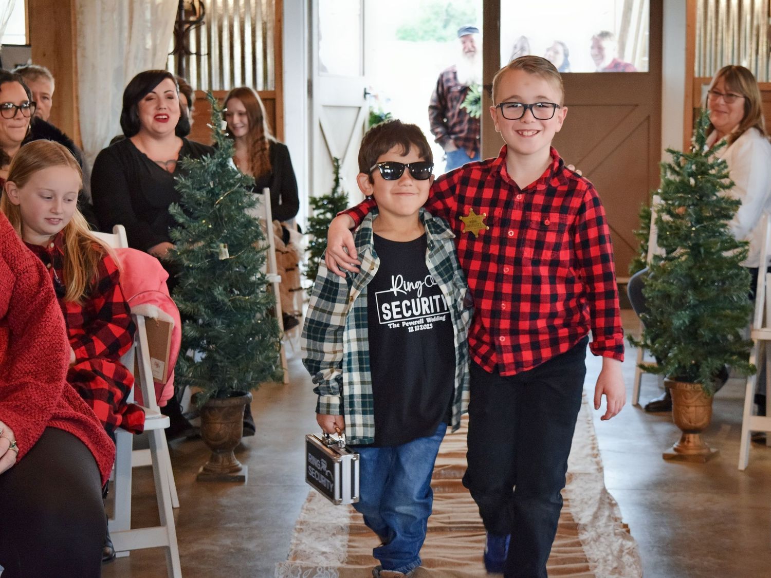 Two young boys walking down wedding aisle in plaid shirts past seated guests and Christmas trees.