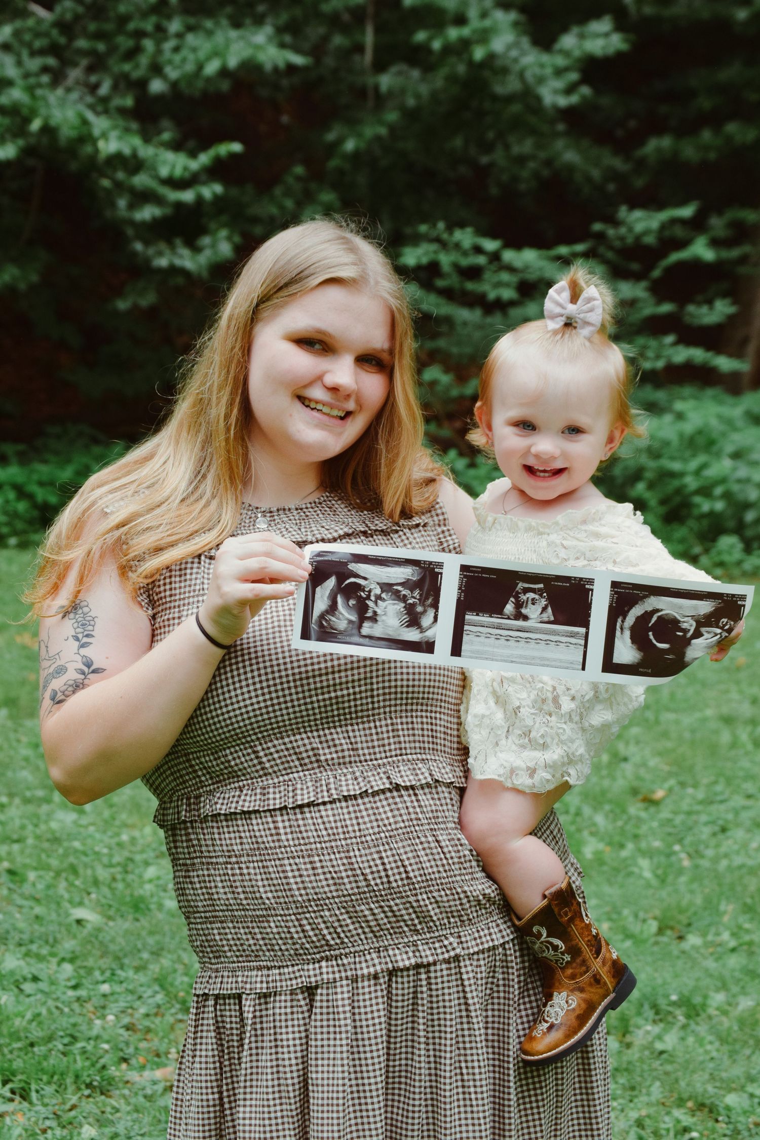 Smiling woman holding toddler girl and ultrasound photos outdoors, announcing pregnancy surrounded by lush green trees.